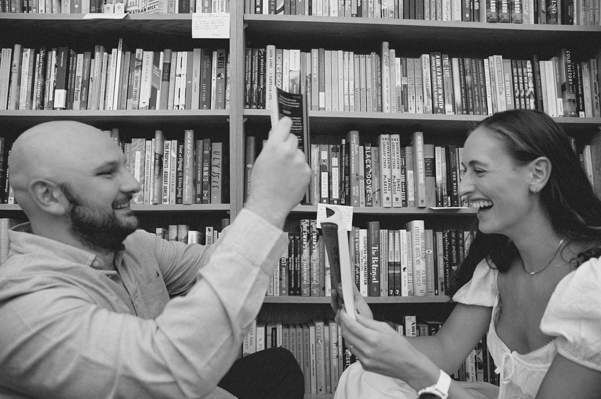 A man and woman laughing and holding cards in front of a wall of books.