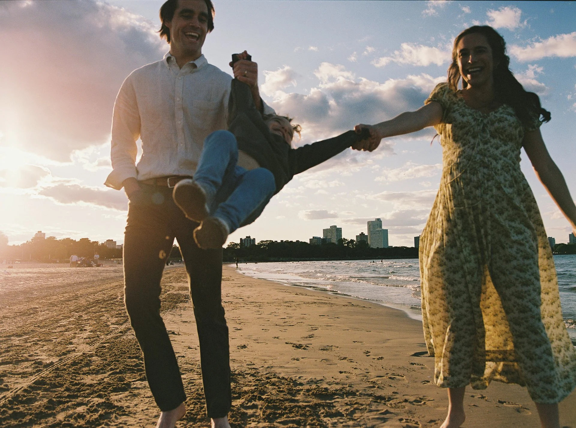 A family of three on the beach during sunset, with a man lifting a young girl and a woman holding her hand as they all smile.