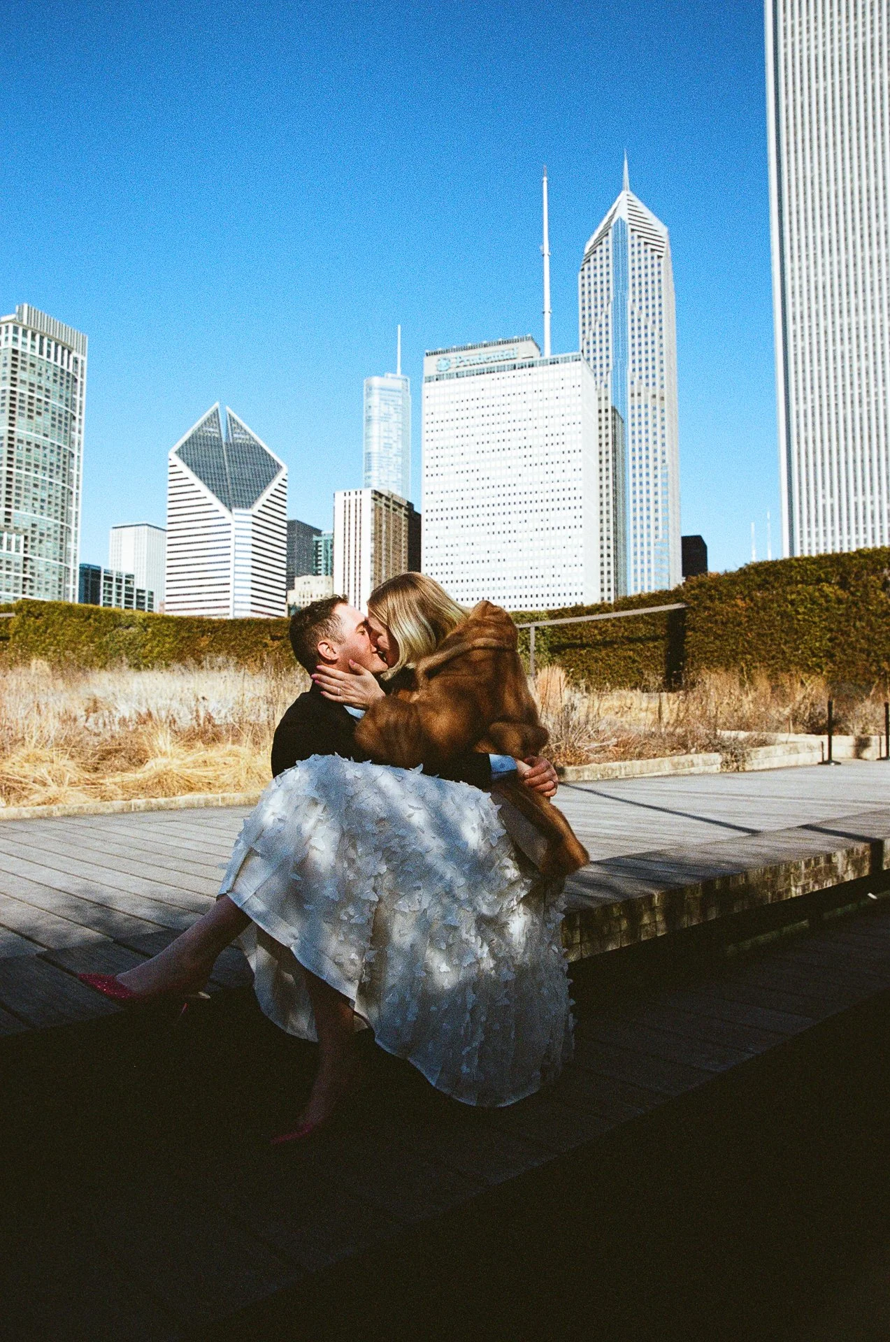 A couple, sitting on a wooden platform, sharing a kiss, while holding a large brown dog, with the city skyline featuring skyscrapers behind them on a sunny day.