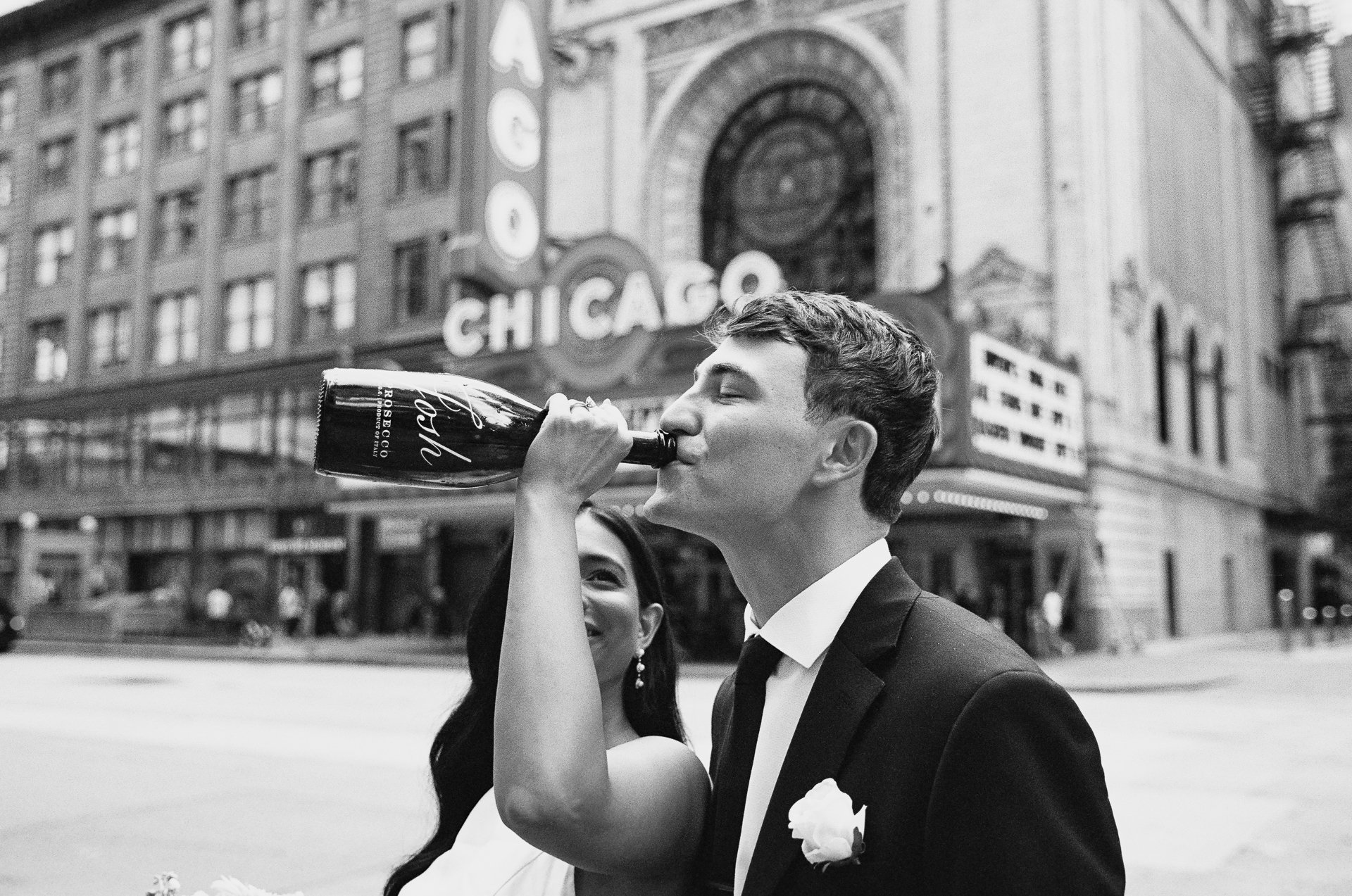 A woman in a wedding dress and earrings is smiling while holding a bottle of Moët & Chandon champagne, pouring it into a man's mouth. The man is wearing a tuxedo with a white flower boutonniere, and they are standing on a city street in front of the 
