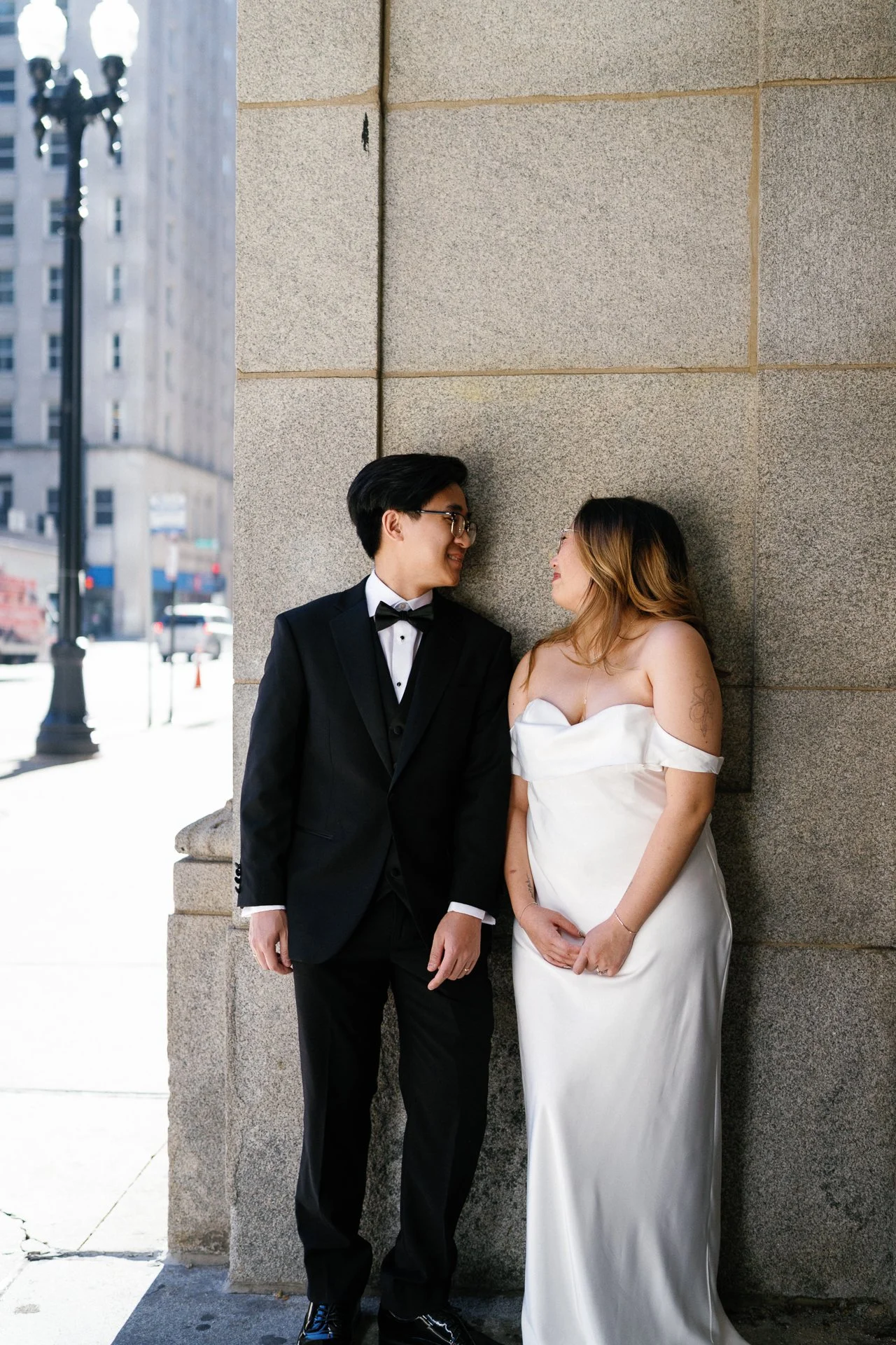 A couple dressed in wedding attire standing against a stone wall on a city street. The man is wearing a black tuxedo with a bow tie, and the woman is in a white, off-the-shoulder wedding gown. They are smiling and looking at each other.