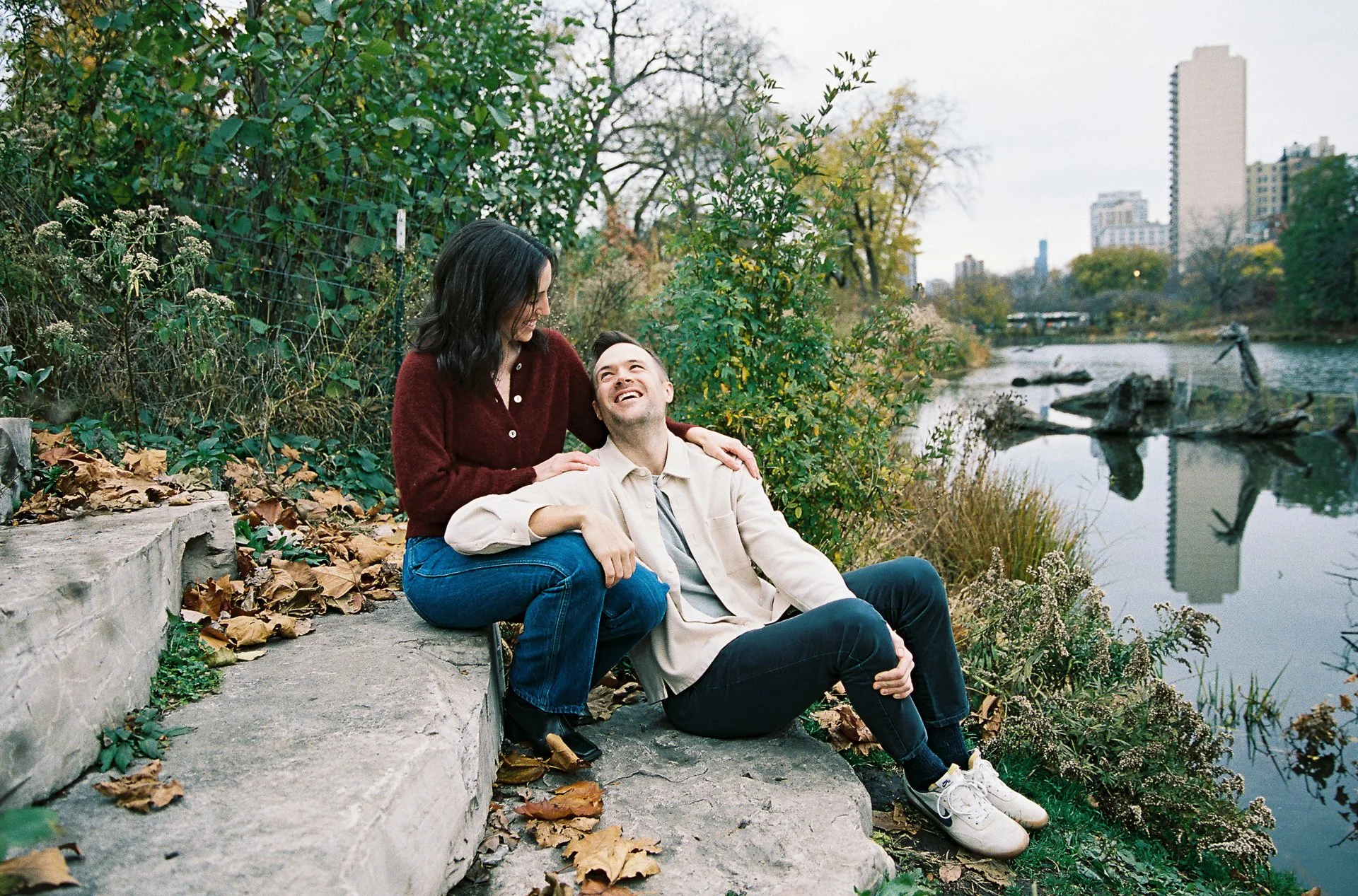 A couple sitting by a river in a park, smiling and looking at each other amidst fall foliage and trees with a city skyline in the background.