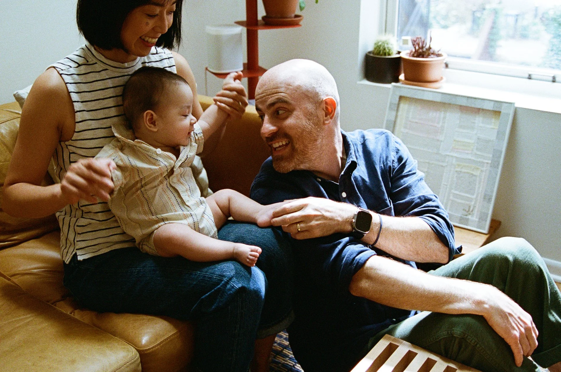 Family sitting on a couch at home, playing and smiling together, with a window and houseplants in the background.