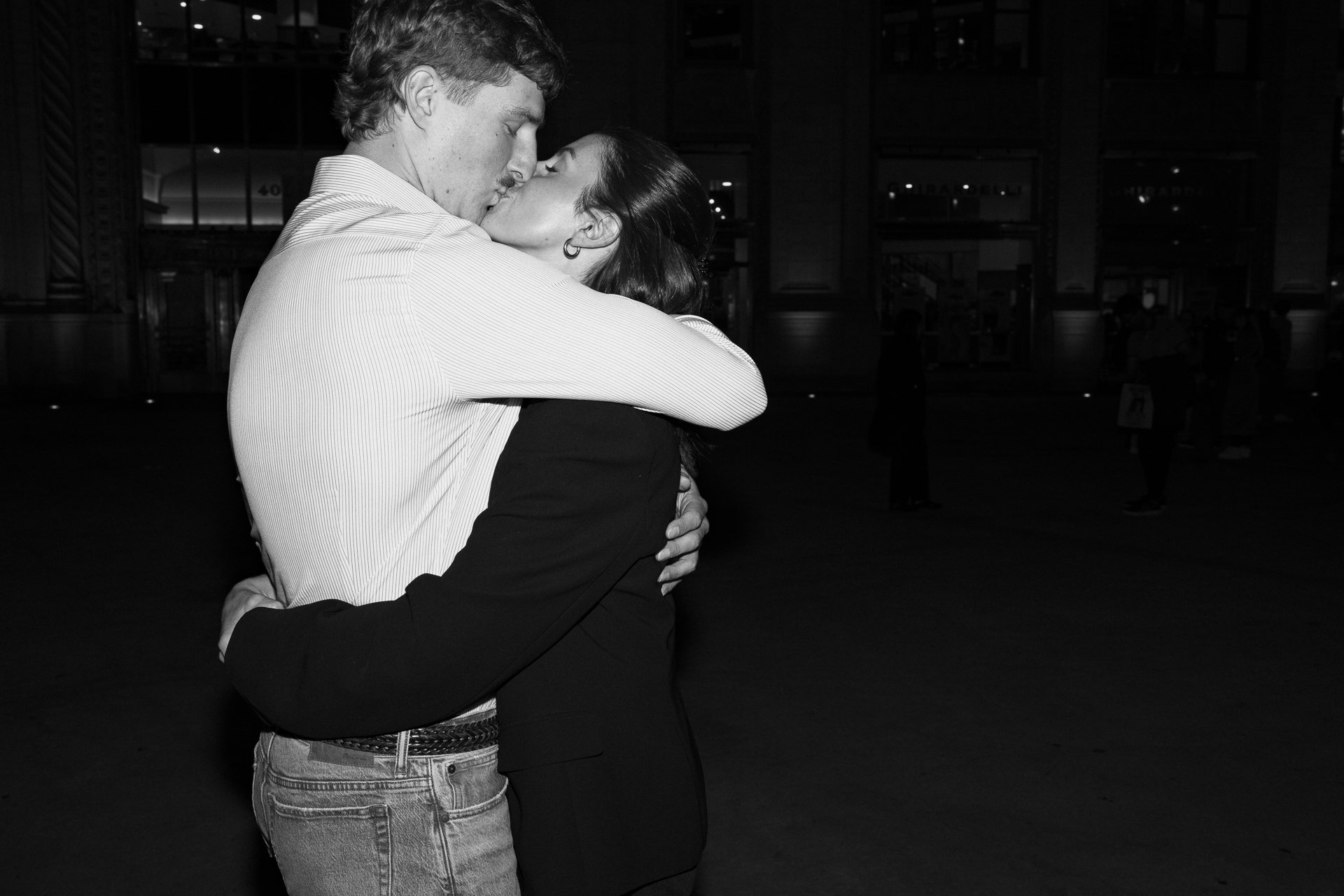 Black and white photo of a young man and woman embracing and kissing, with a dark background and a few people visible in the distance.