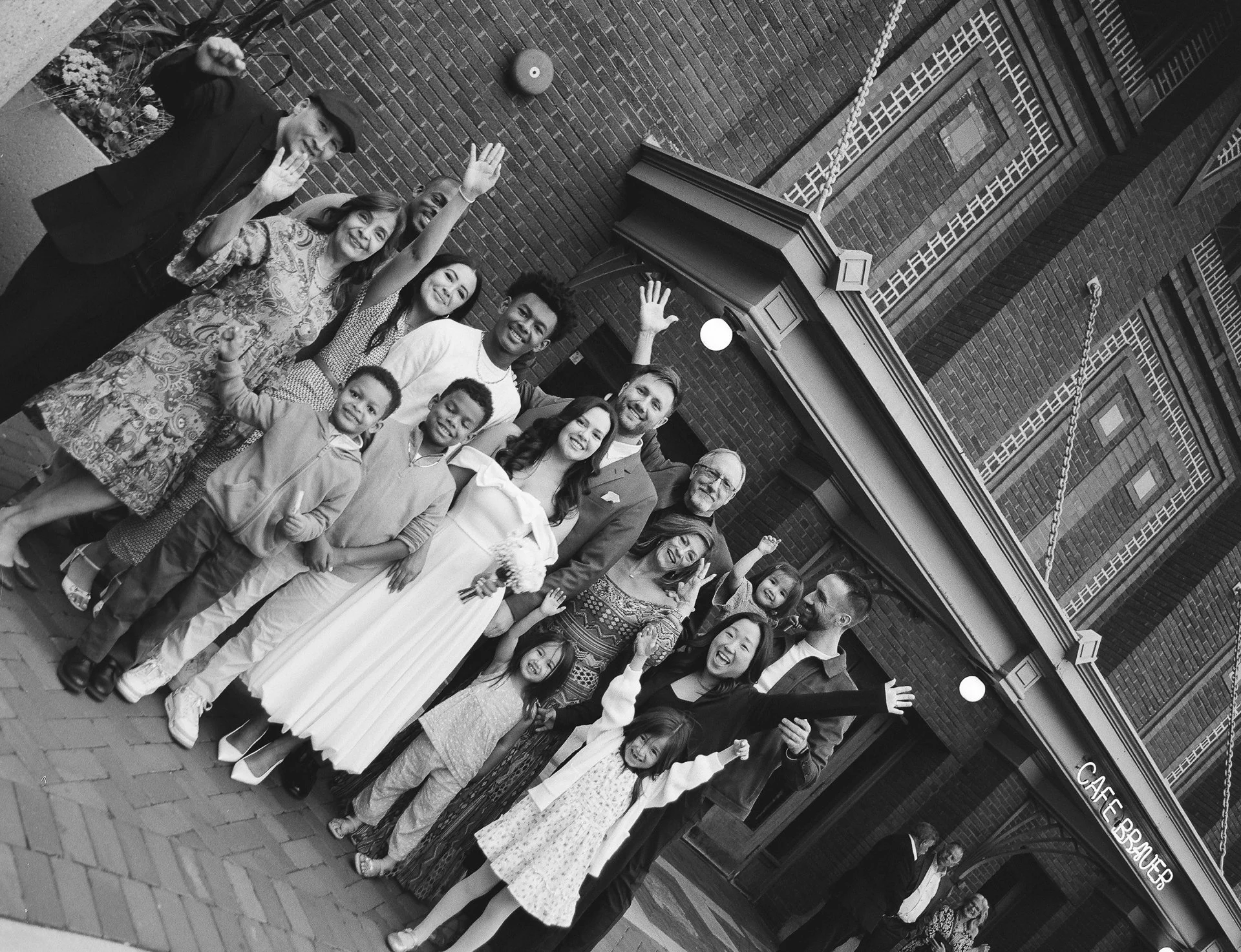 Group of diverse people, including children and adults, standing together outside a brick building with a sign that reads 'Cafe Brauers', smiling and waving at the camera.