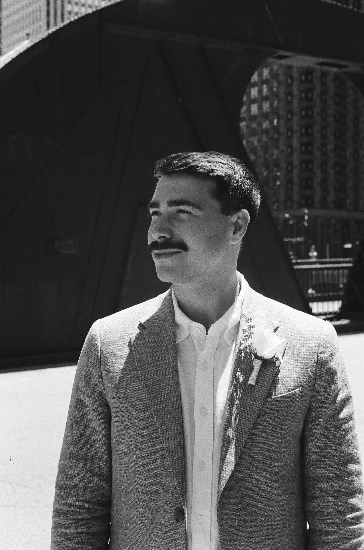 A black-and-white photo of a man with a mustache wearing a light-colored suit and a white shirt, standing outdoors with a modern building structure in the background.