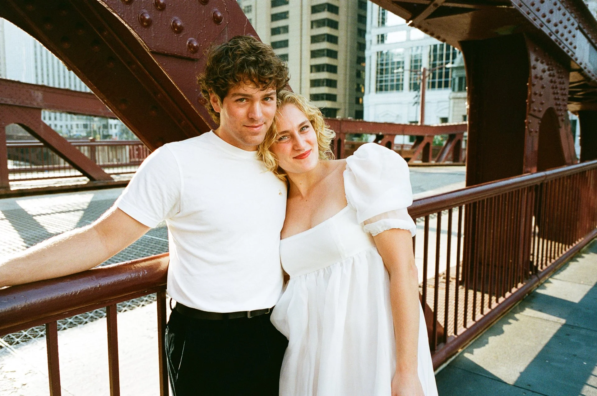 A young man and woman standing close together on a bridge in an urban city setting, with the man wearing a white t-shirt and the woman wearing a white dress with puffed sleeves, both smiling gently at the camera.