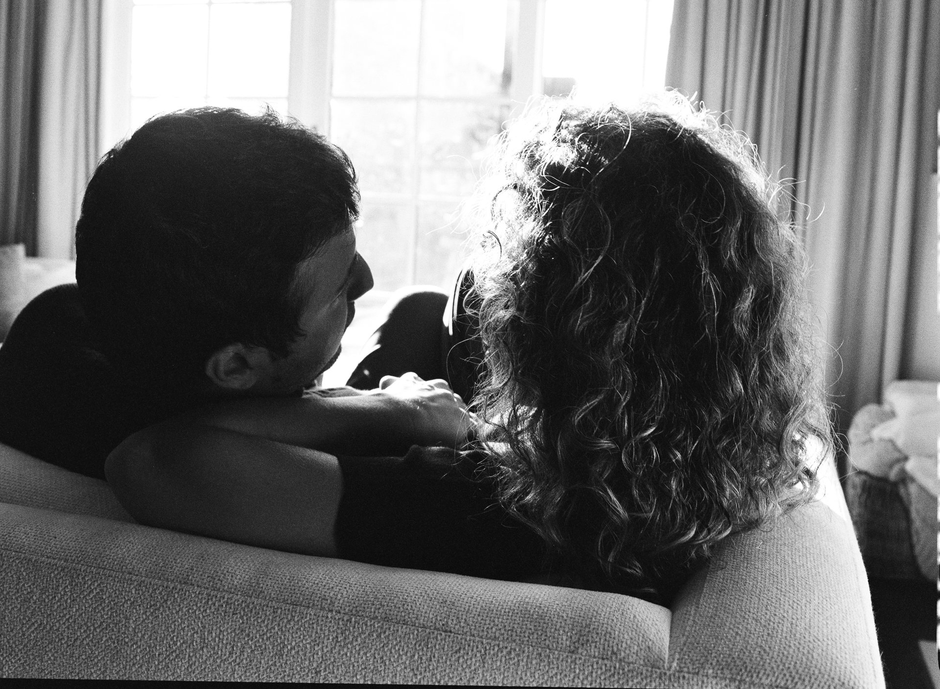 A man and a woman sitting on a couch in front of a window, with the man leaning on the woman's arm.