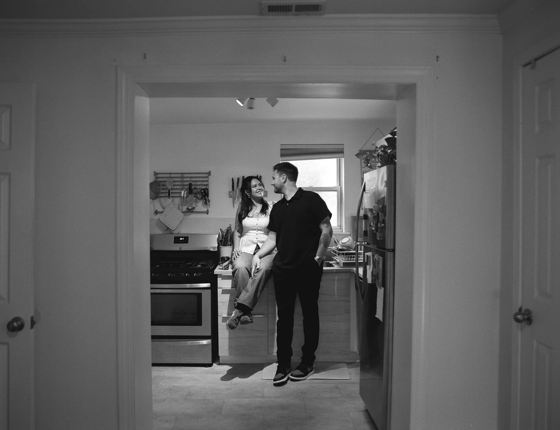 A man and woman sharing a moment in a kitchen, with the woman sitting on the counter and the man standing. Black and white photograph.