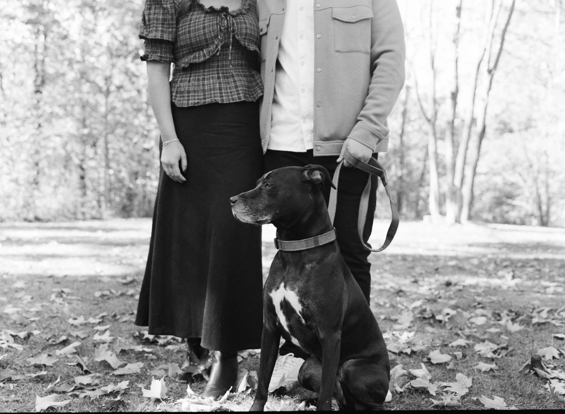 A black and white photo of a person standing outdoors in a park with a large dog sitting in front of them. The person is wearing a long skirt and a jacket, and the dog is on a leash, looking to the side.