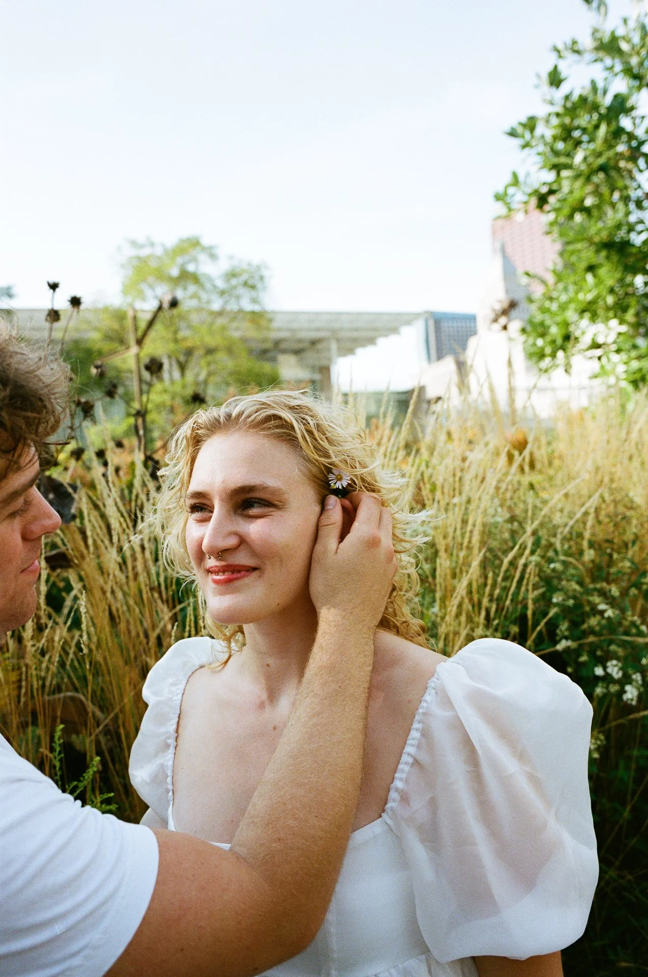 A person adjusting a woman's earring outdoors among tall grass and plants.