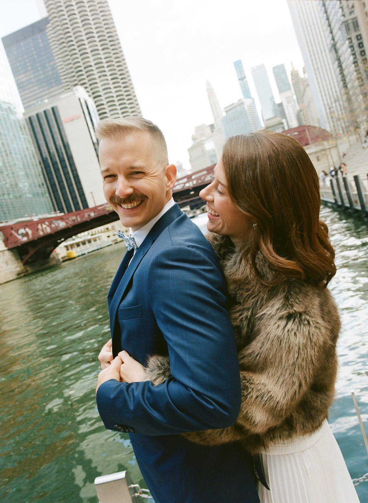 A smiling man in a blue suit and a woman in a fur coat standing by the river with city buildings in the background.