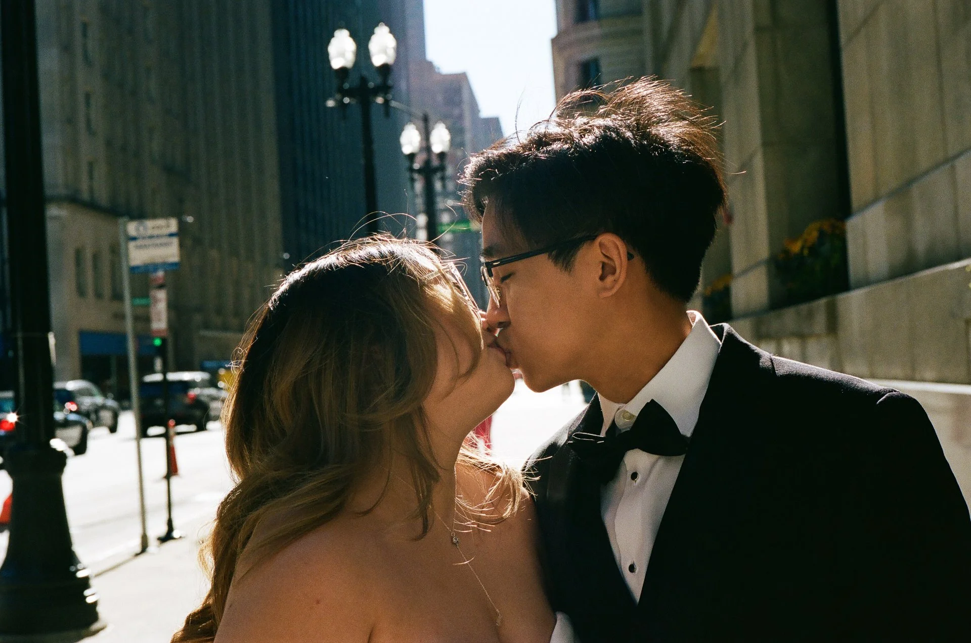 A couple kissing on a city street during daytime, with the man wearing a tuxedo and the woman wearing a strapless dress.