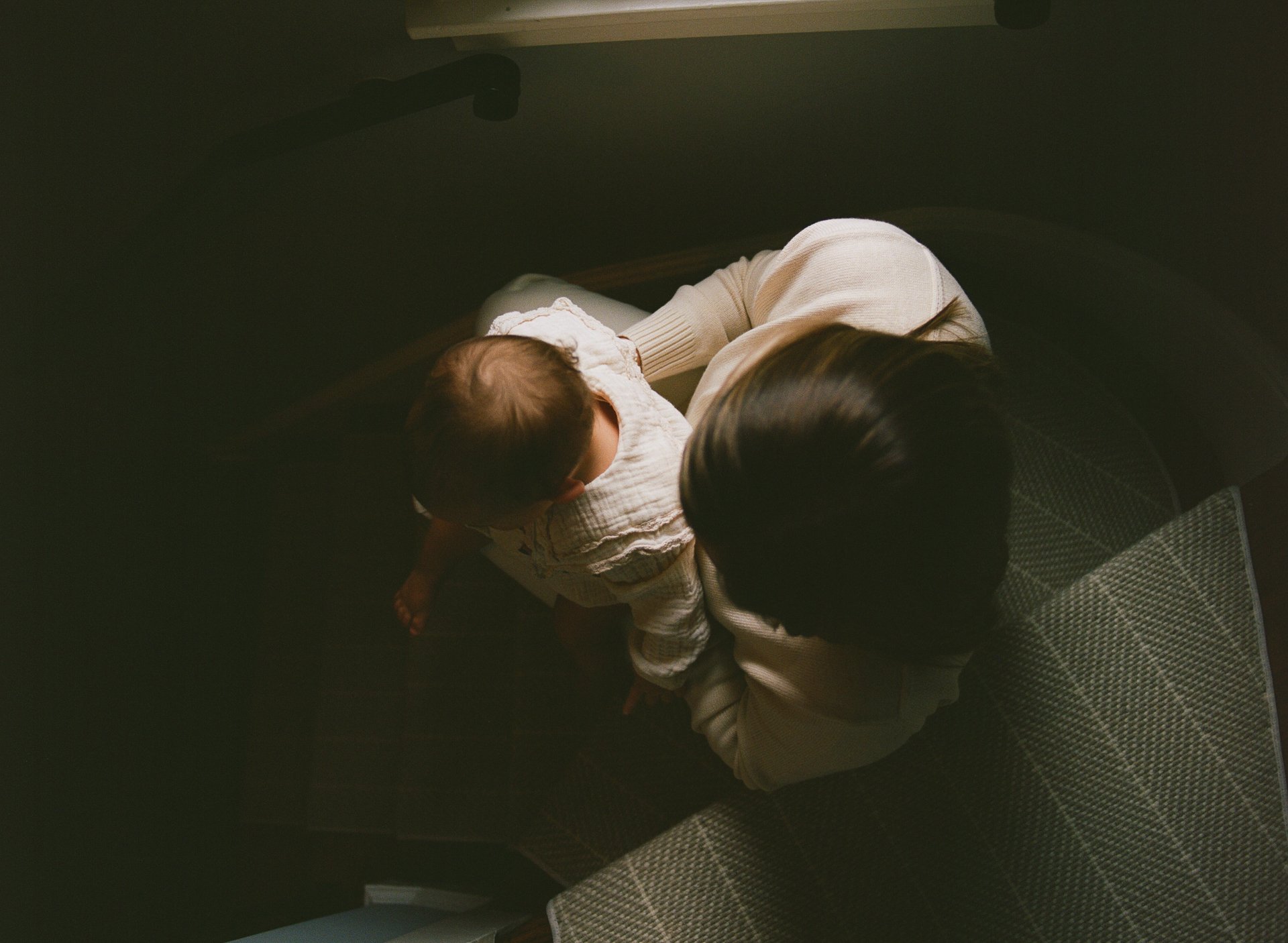 An adult holding a baby on their lap, seen from above, in a dimly lit room.
