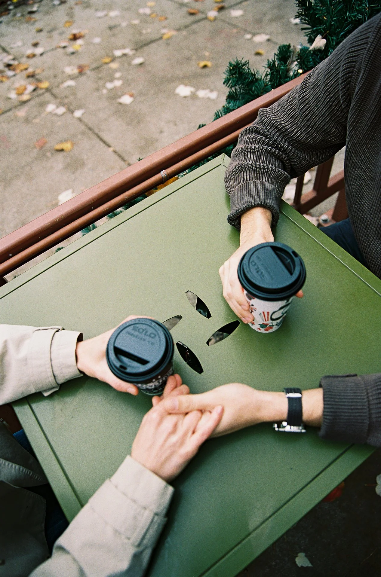 Two people sitting at a green metal table outdoors, holding disposable coffee cups, with some hands clasped on the table.