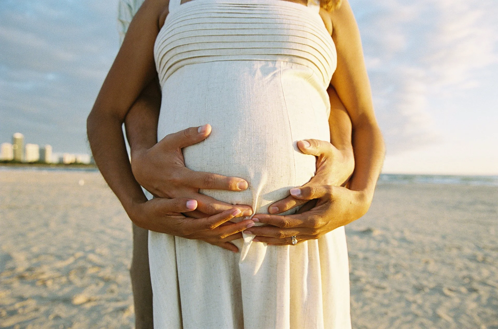 A pregnant woman in a white dress on the beach, with her hands gently resting on her belly, surrounded by another person’s hands.