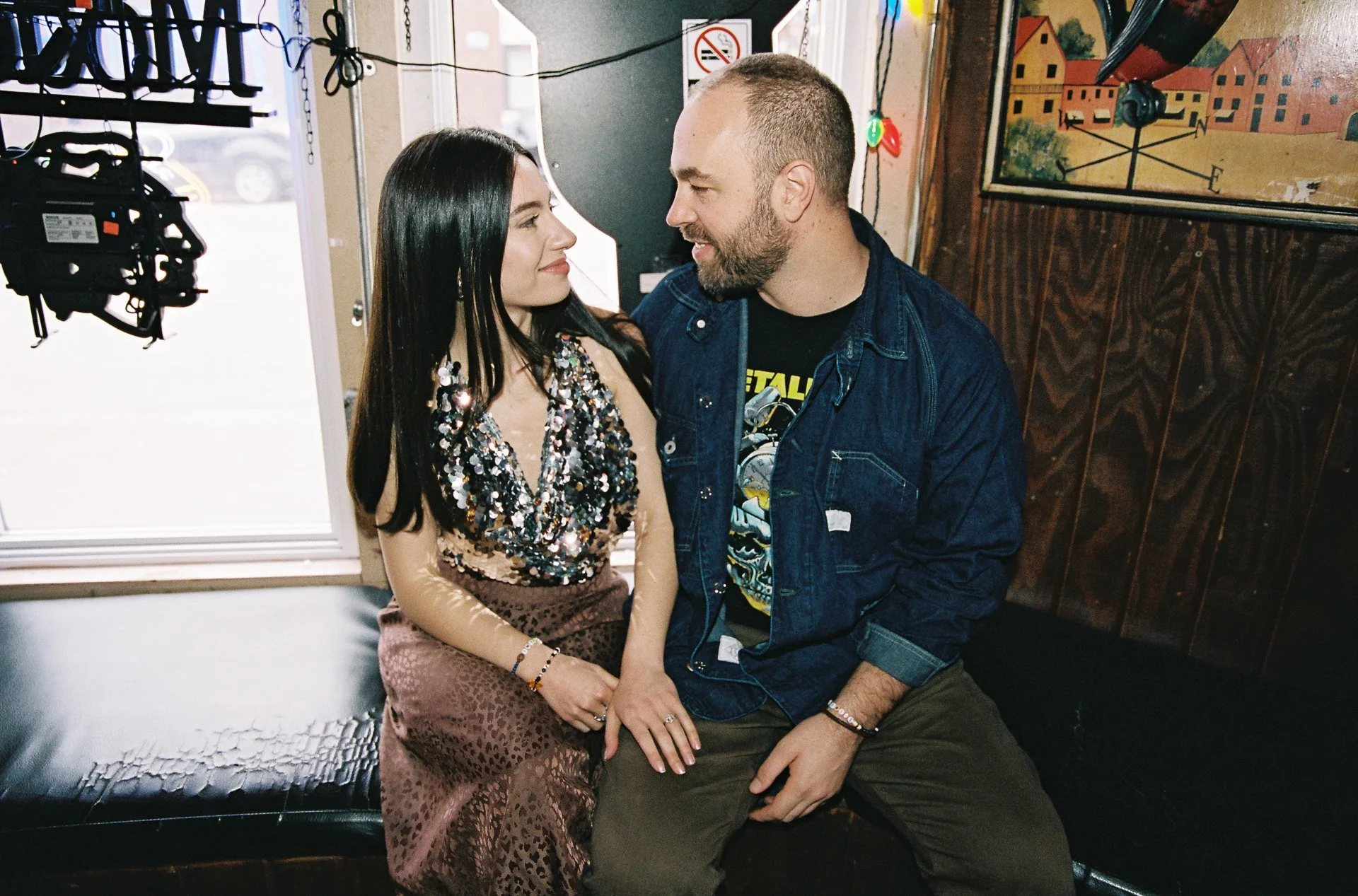 A woman with long black hair wearing a sequined top and a leopard print skirt sits on a black leather bench. A man with a beard, wearing a denim jacket and a Metallica t-shirt, sits close to her on the same bench, facing each other and smiling. There