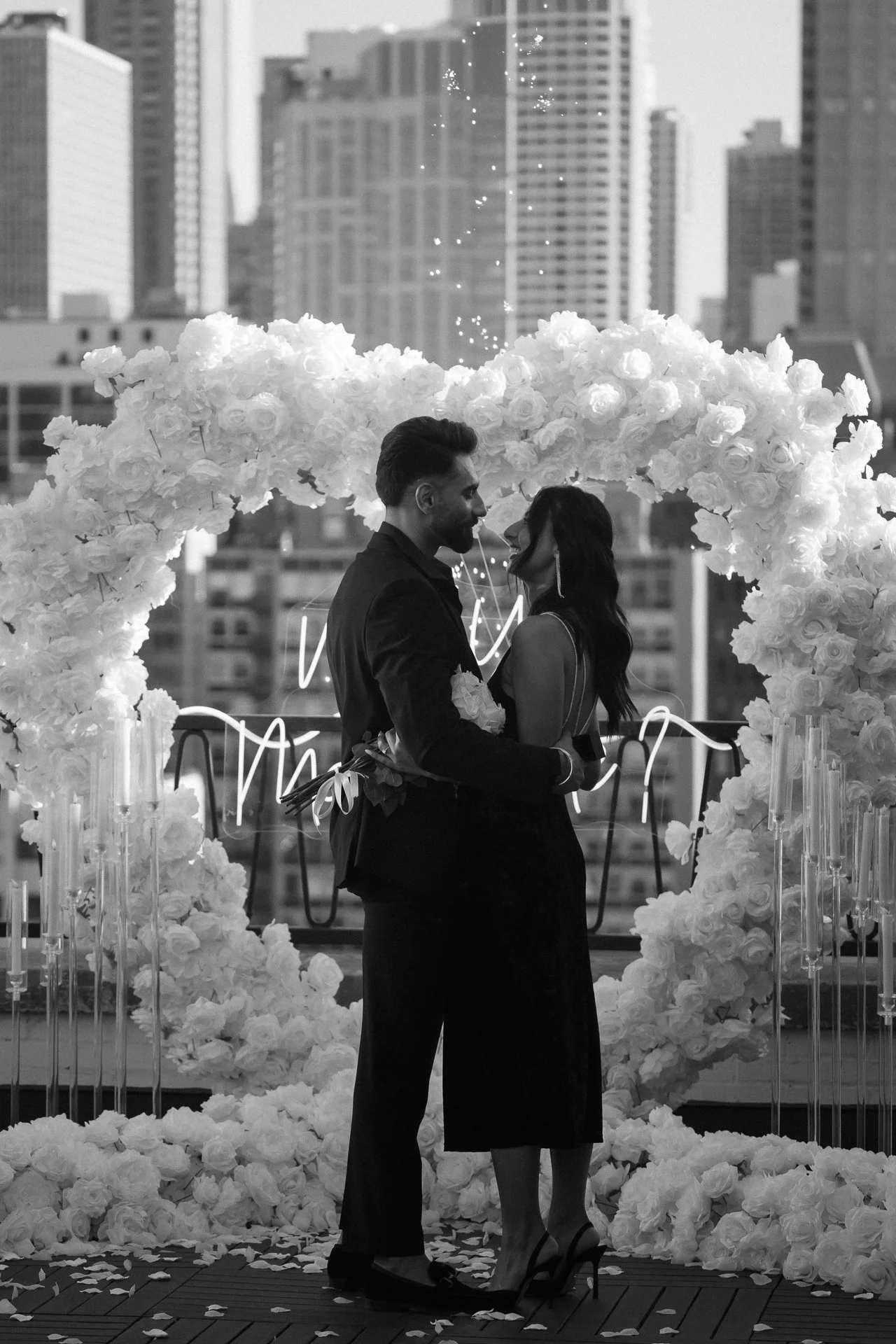 A couple stands close together, holding a bouquet, under a floral arch on a city rooftop, with tall buildings in the background.