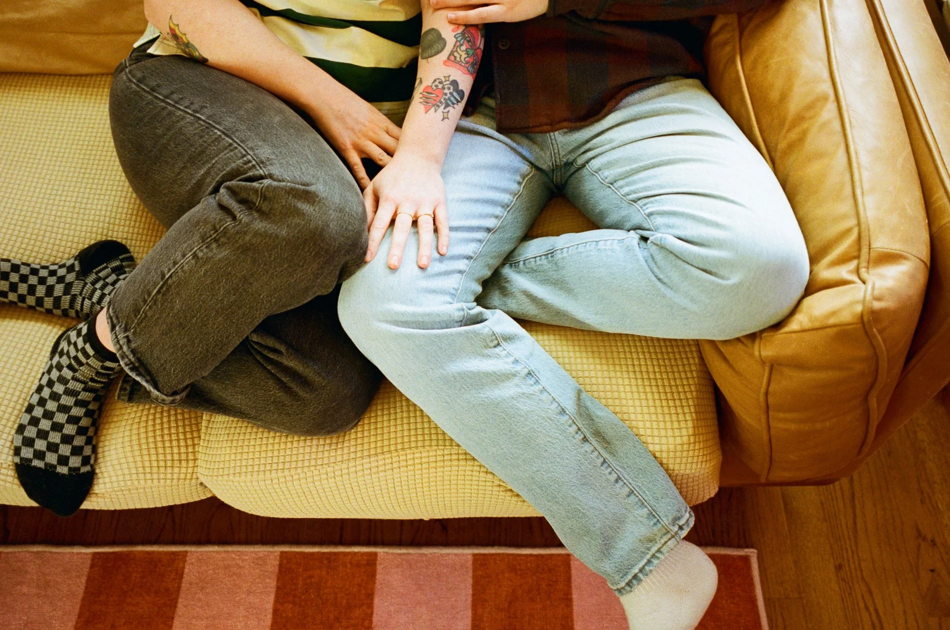 Two people sitting on a sofa with one person's legs intertwined with the other's. They are close, with one person's hand on the other's leg. One person has tattoos on their arm, and both are wearing casual clothes, including jeans and checkered socks