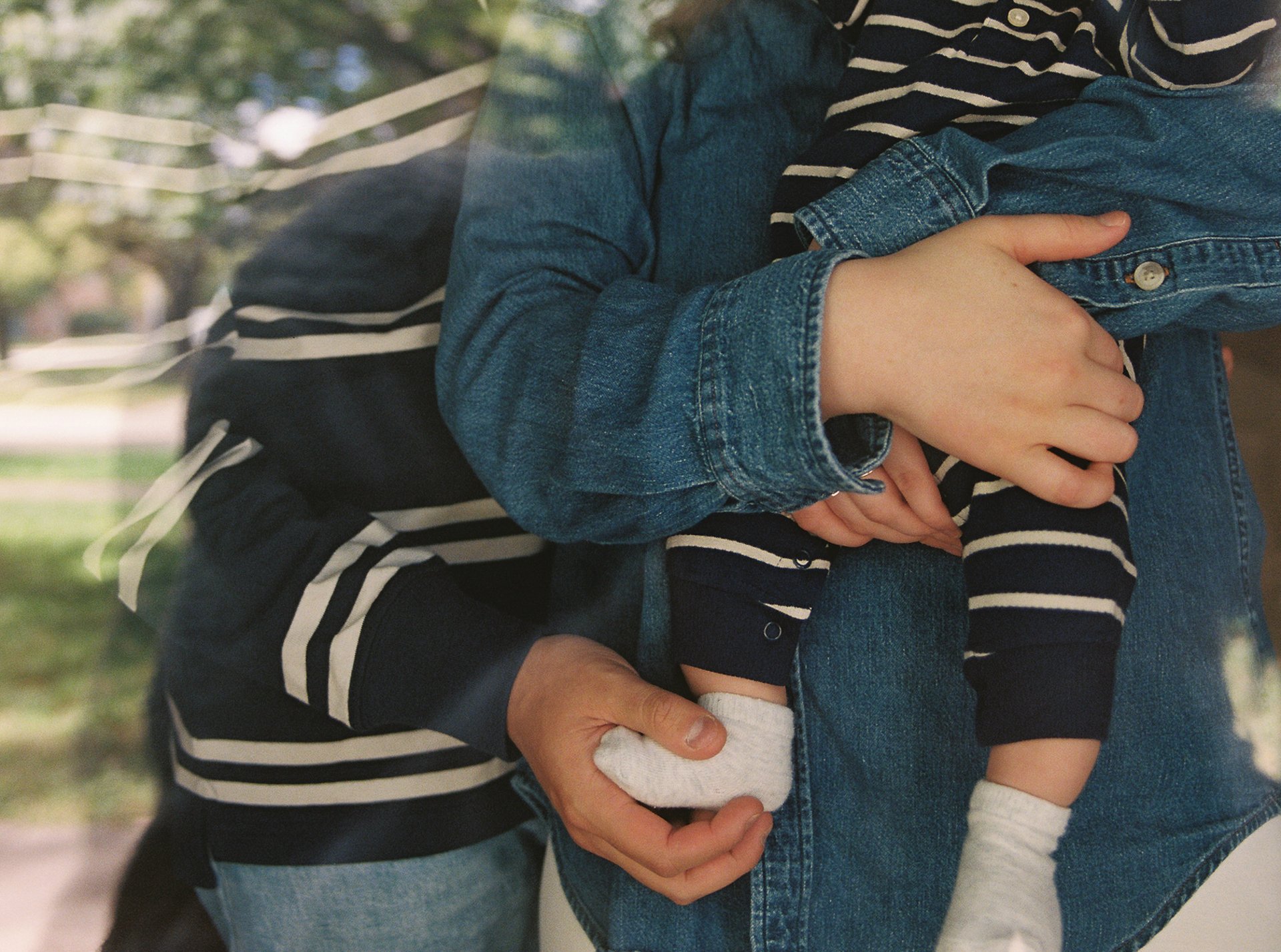 Close-up of an adult holding a child, with their arms crossed. The adult is wearing a denim jacket and jeans, while the child is dressed in a striped shirt, navy pants, and white socks, with their legs resting on the adult's arm.