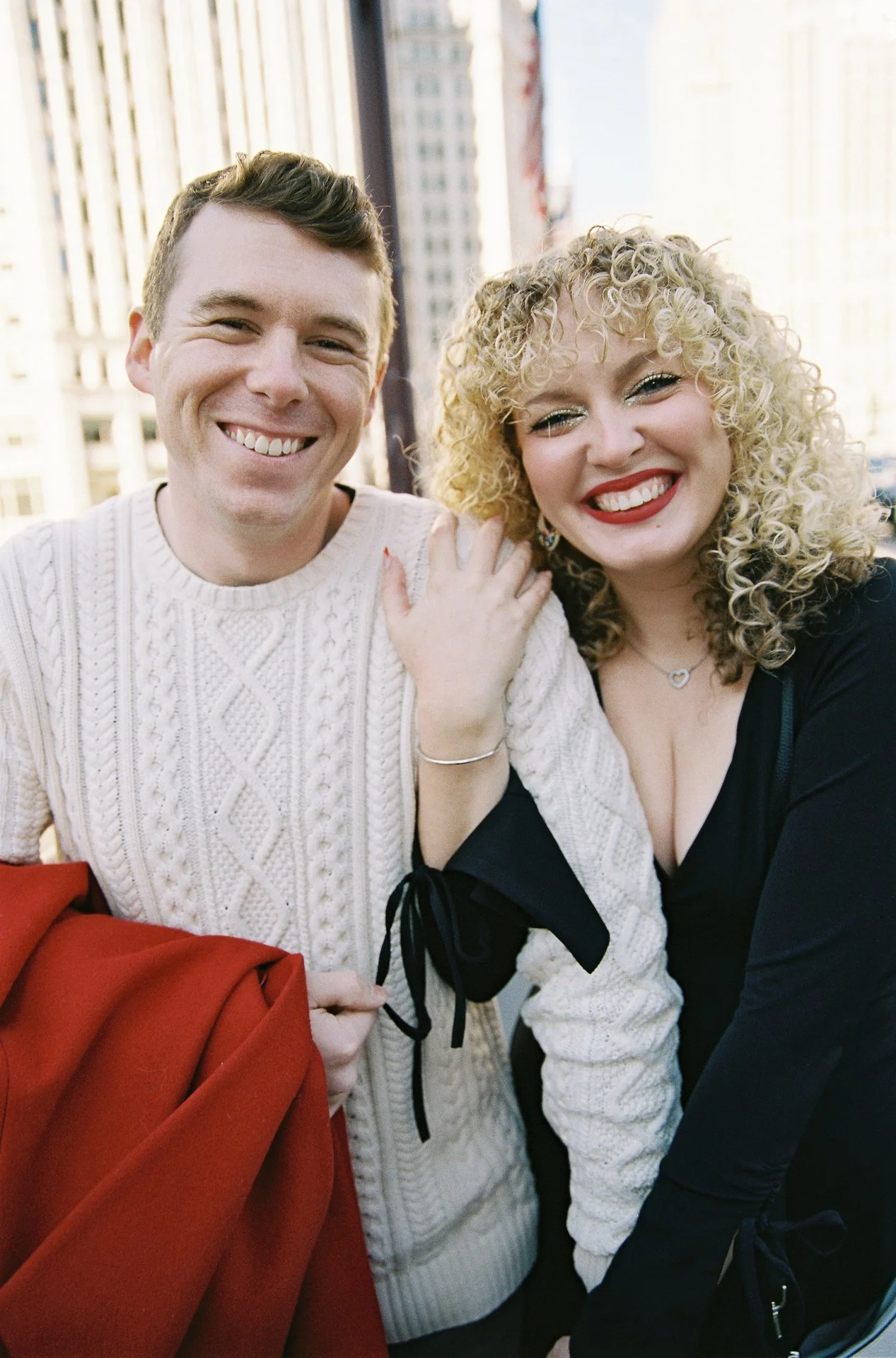 Two smiling people, a man and a woman, close together with city buildings in the background.