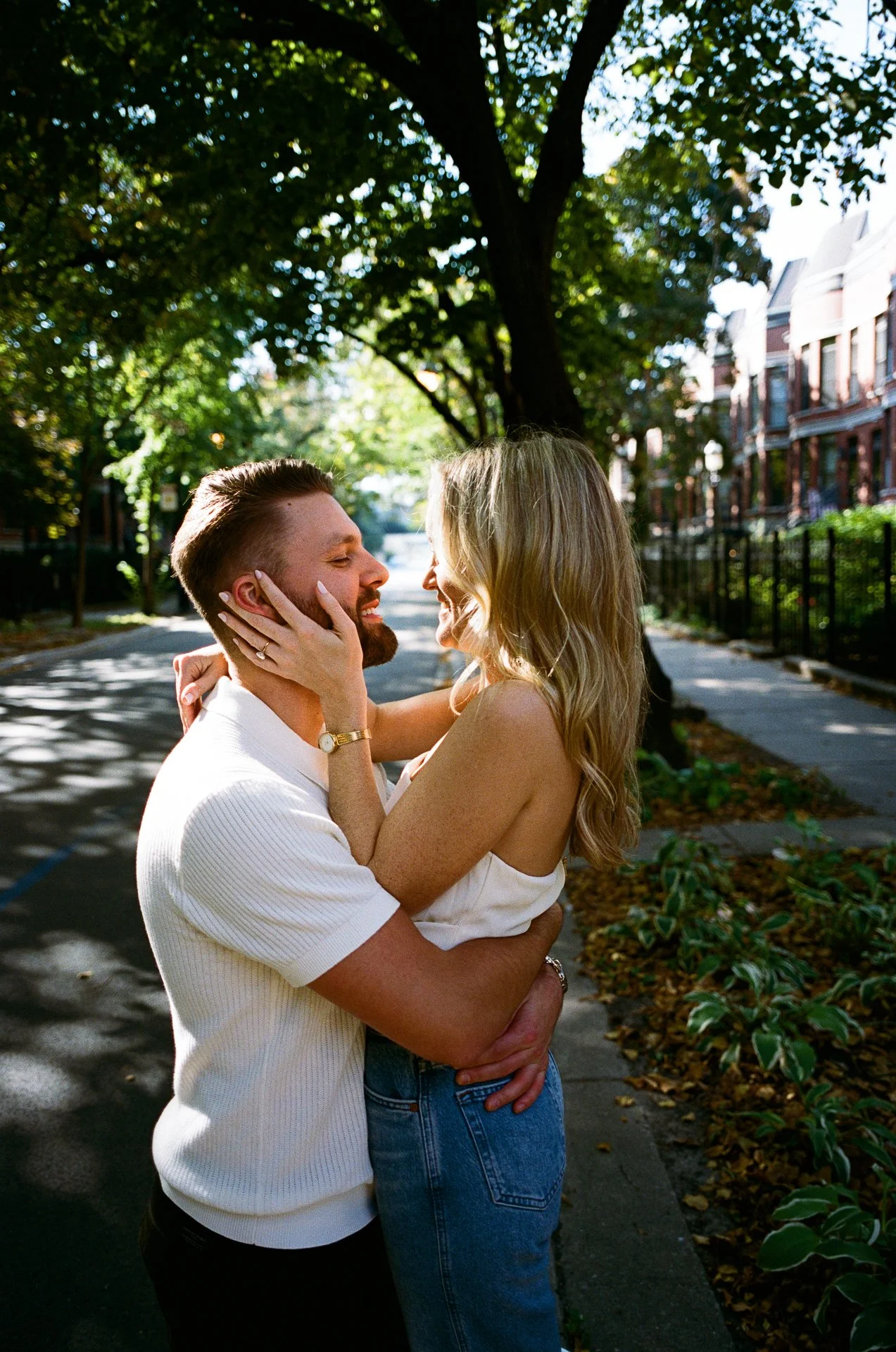 A smiling couple embracing on a tree-lined sidewalk during daytime, with trees and residential buildings in the background.