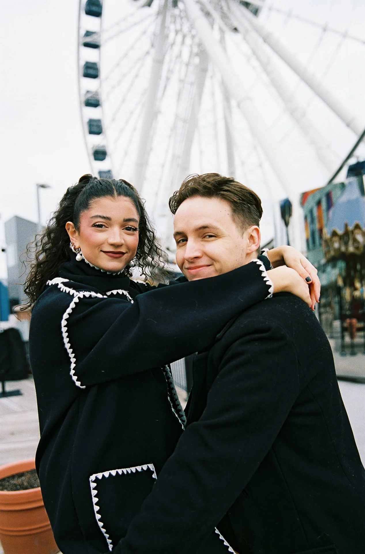 A young woman and a young man are embracing in front of a large Ferris wheel at a carnival or amusement park. The woman has curly dark hair and is wearing a black jacket with white trim, and the man has short brown hair and is wearing a black jacket.