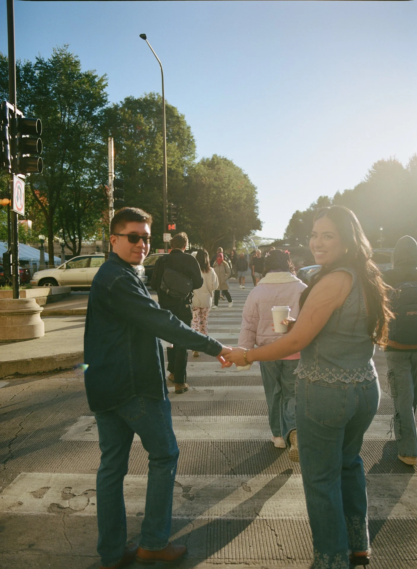 A young man and woman holding hands on a city crosswalk during daylight, with other pedestrians and cars in the background, trees, and traffic lights.