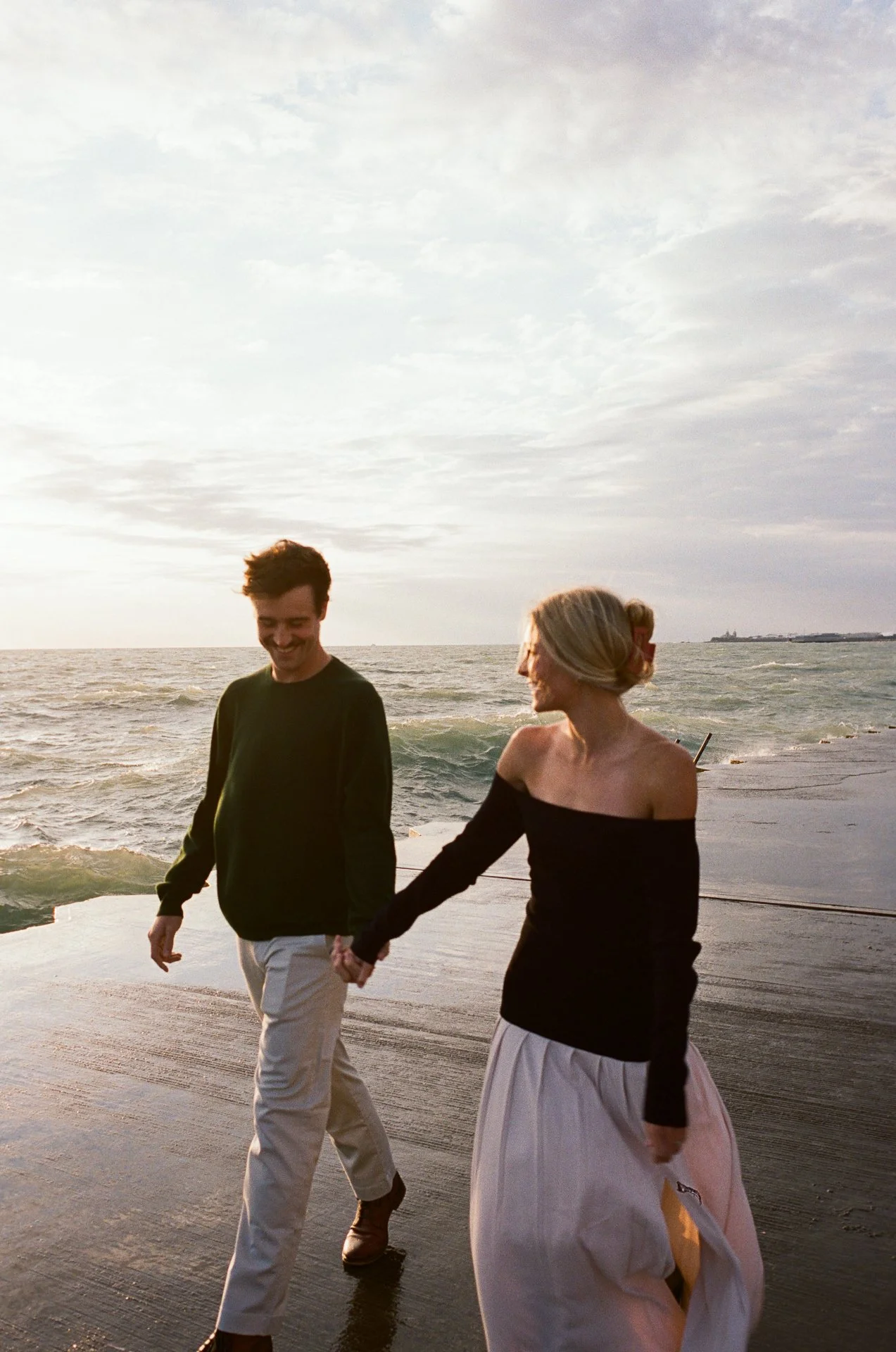 A young man and woman walk hand in hand along a beach walkway during sunset, with the ocean waves in the background.