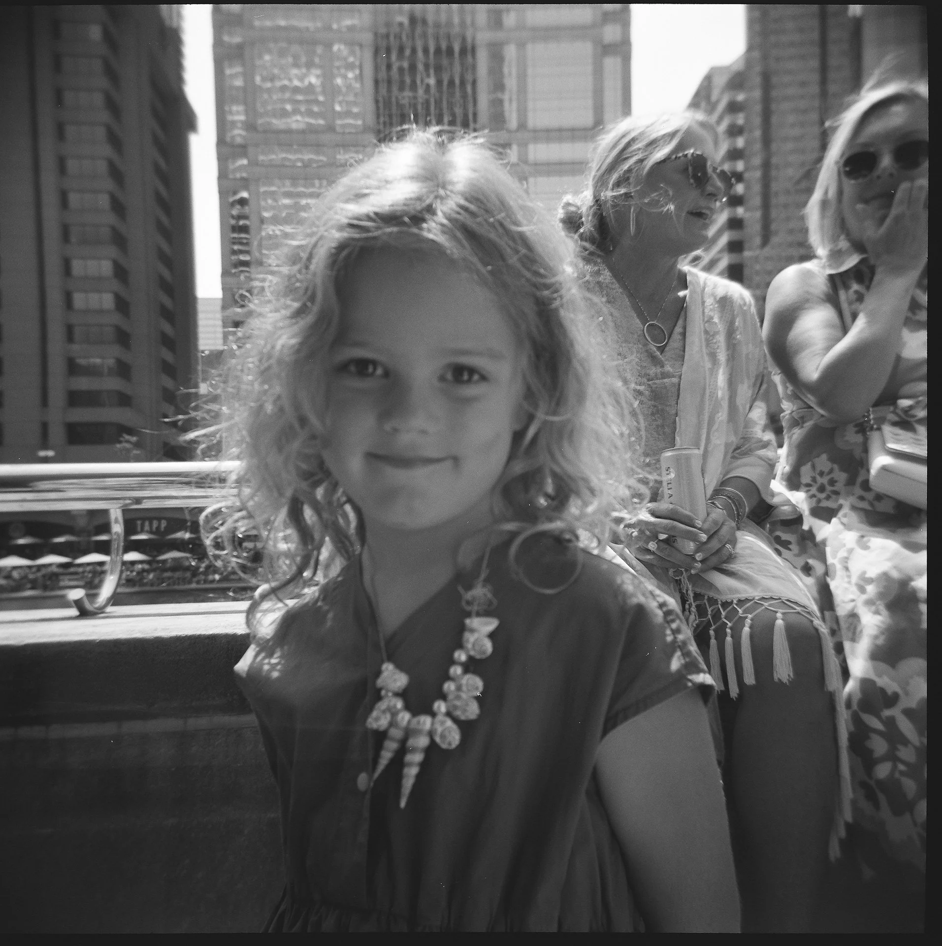 A young girl with curly hair wearing a necklace and smiling at the camera, seated outdoors on a city street with tall buildings and two women in the background.
