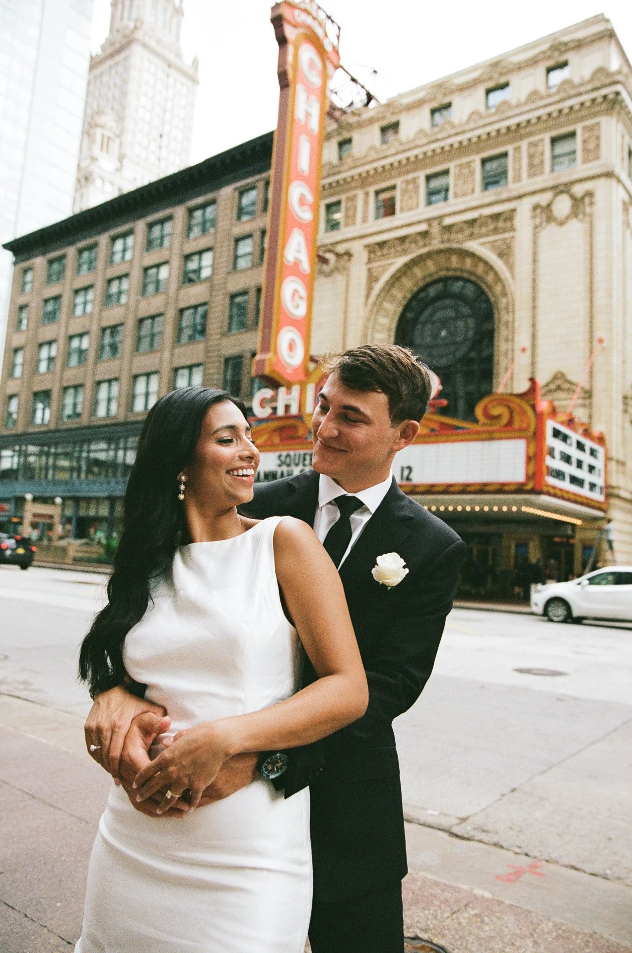 A smiling bride in a white dress and a groom in a black suit with a tie, standing on a city street in front of a theater marquee that reads 'Chicago'.