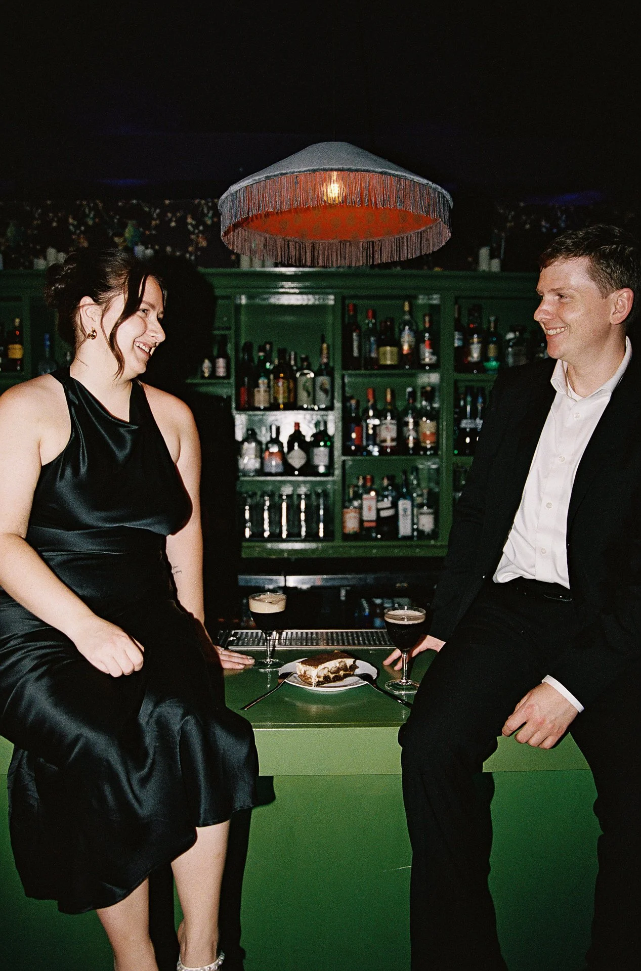 A woman and a man sitting on a green bar with a cake and two drinks, smiling at each other, in a dimly lit bar.