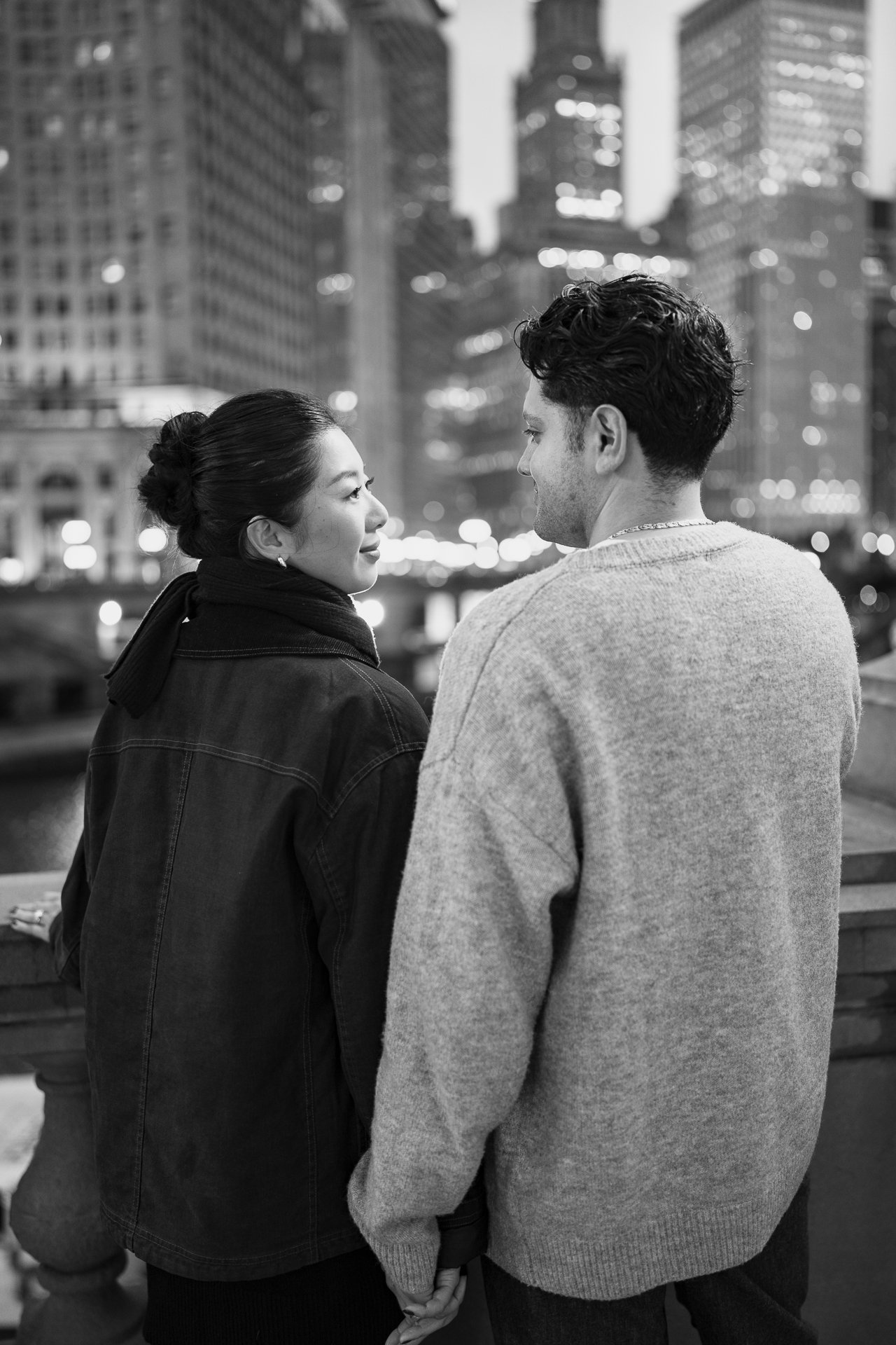 A black-and-white photo of a couple holding hands and looking into each other's eyes against an urban cityscape at night with tall buildings and blurred city lights in the background.