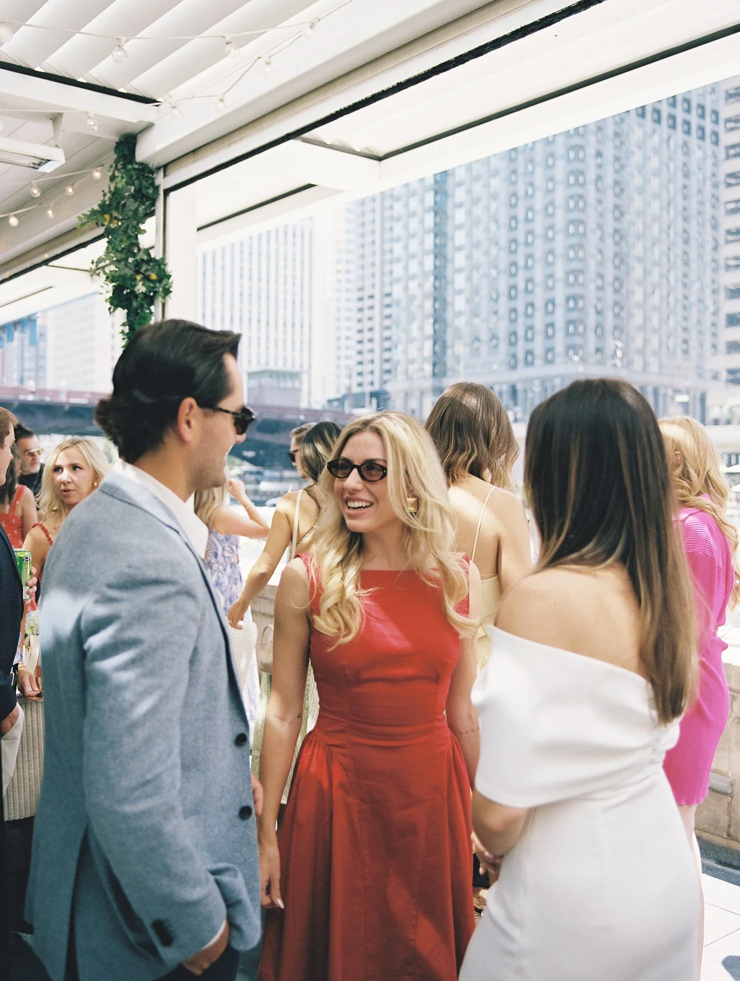 People socializing on a rooftop terrace in a city with tall buildings, including a woman in a red dress and two people talking closely in the foreground.