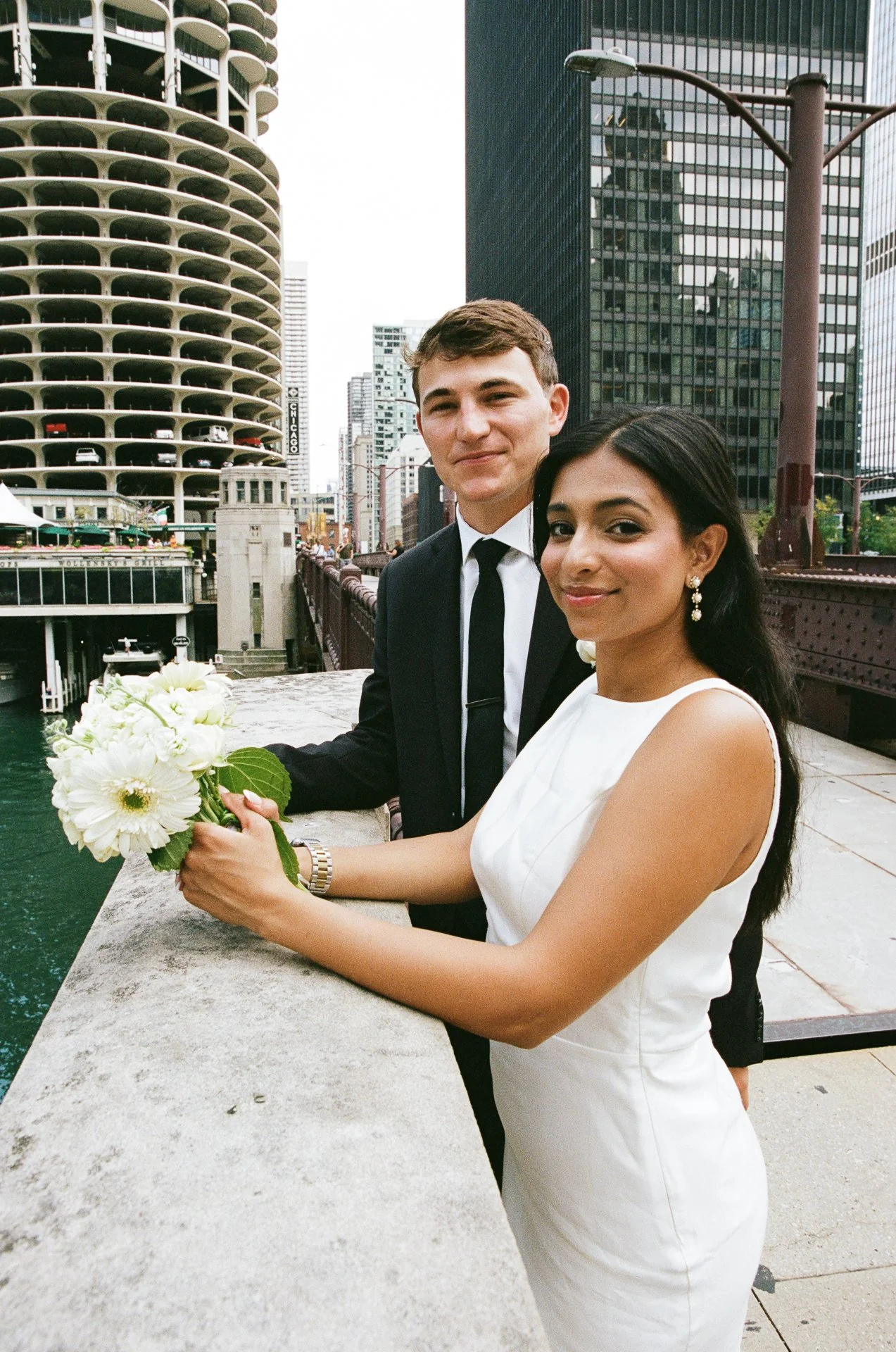 A bride and groom standing on a city bridge with tall buildings in the background, the bride holding a bouquet of white flowers, both smiling.