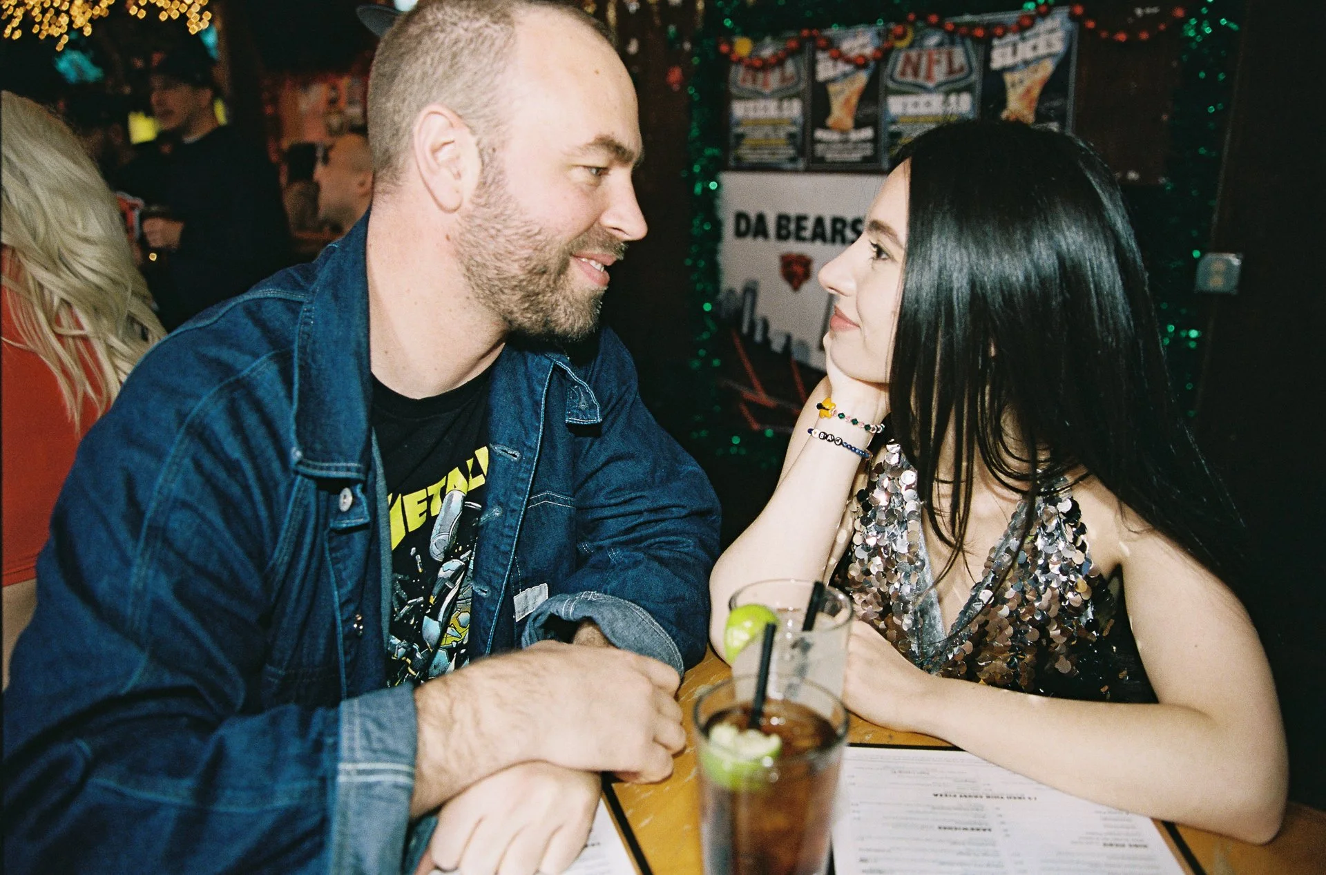 A man and a woman sitting at a bar or restaurant, looking at each other and smiling, with drinks in front of them. The background features sports memorabilia and other patrons.