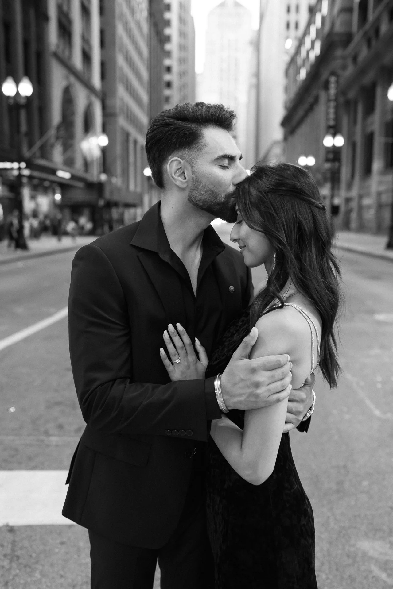 A black-and-white photo of a man and woman embracing on a city street, the man kissing the woman's forehead with eyes closed, both dressed formally.
