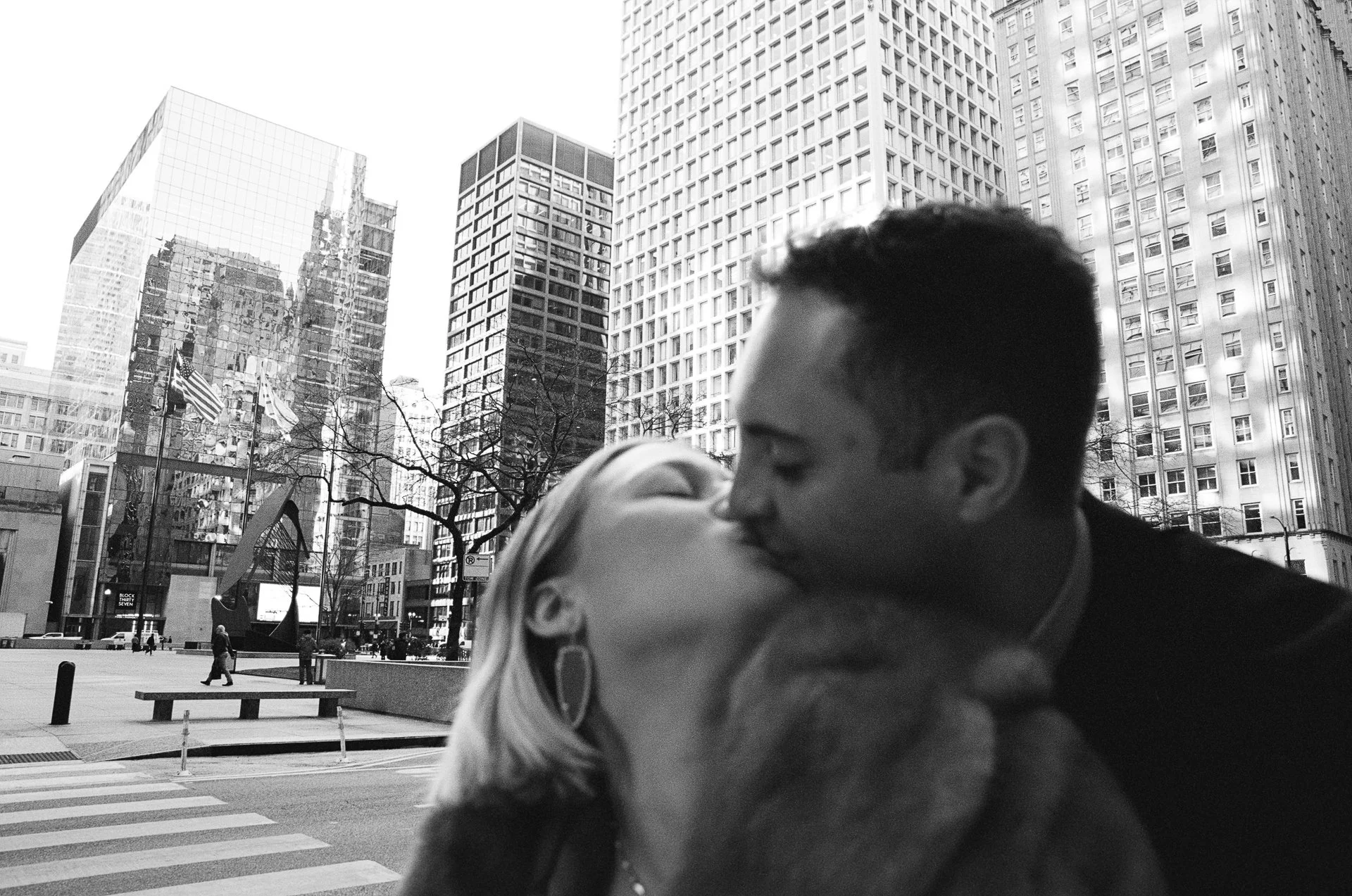 A man and woman kissing in a city street with tall office buildings in the background, black-and-white photograph.