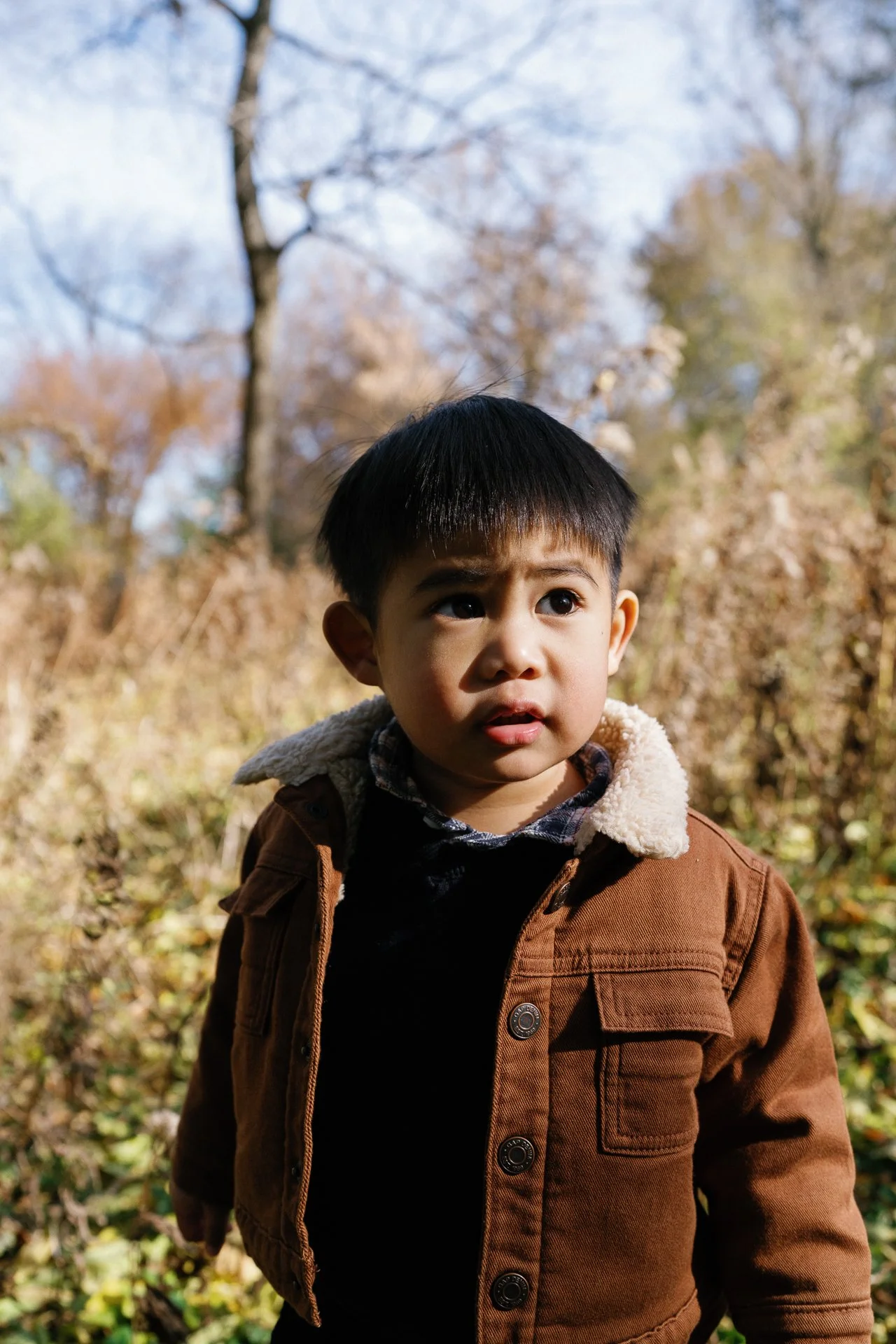 A young boy with dark hair and brown eyes looks to the side outdoors during fall, wearing a brown jacket with a fleece collar, surrounded by trees with autumn foliage.