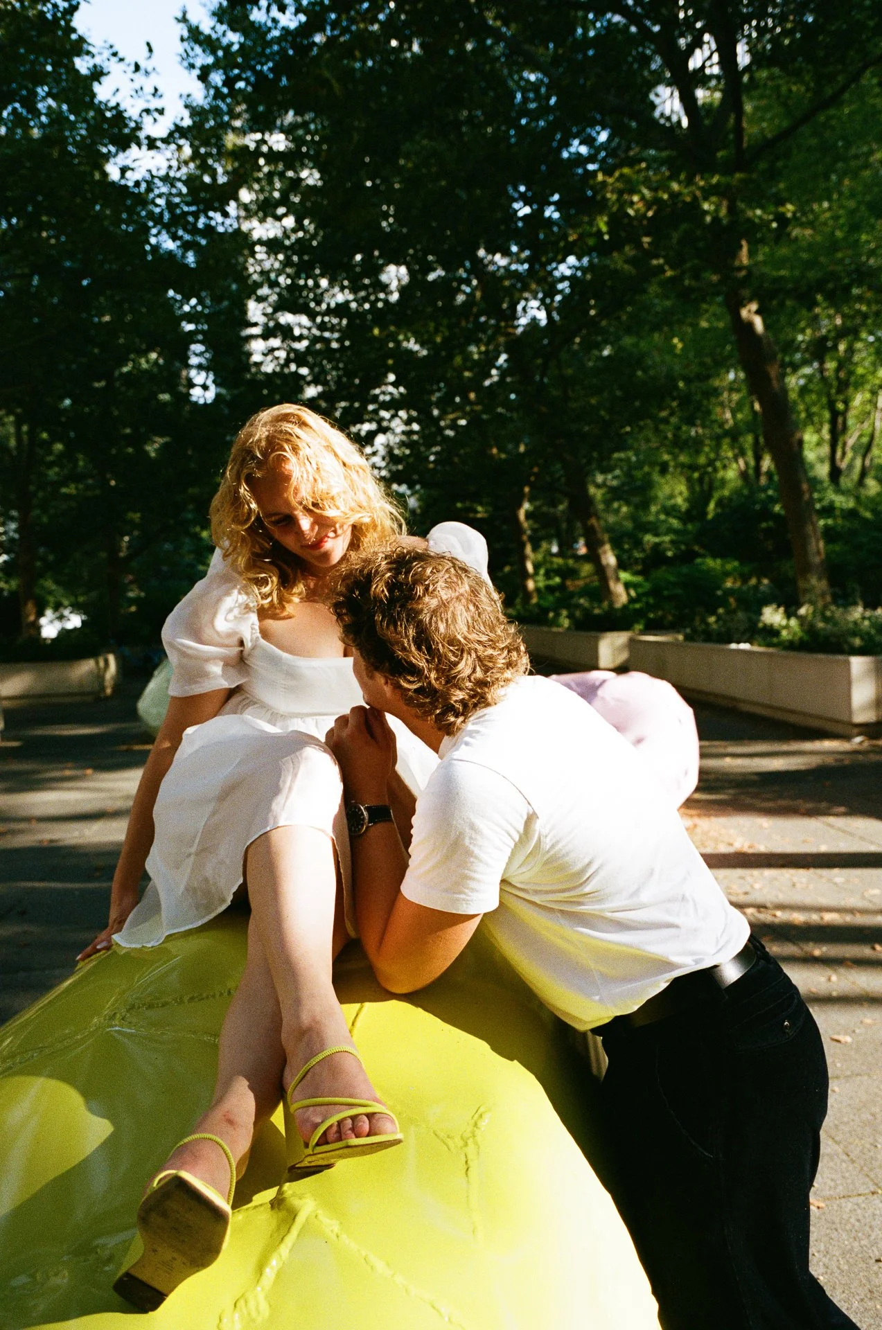 A couple is sitting on a yellow sculpture outdoors in a park with trees, with the woman wearing a white dress and yellow heels, and the man leaning in as they share a tender moment.