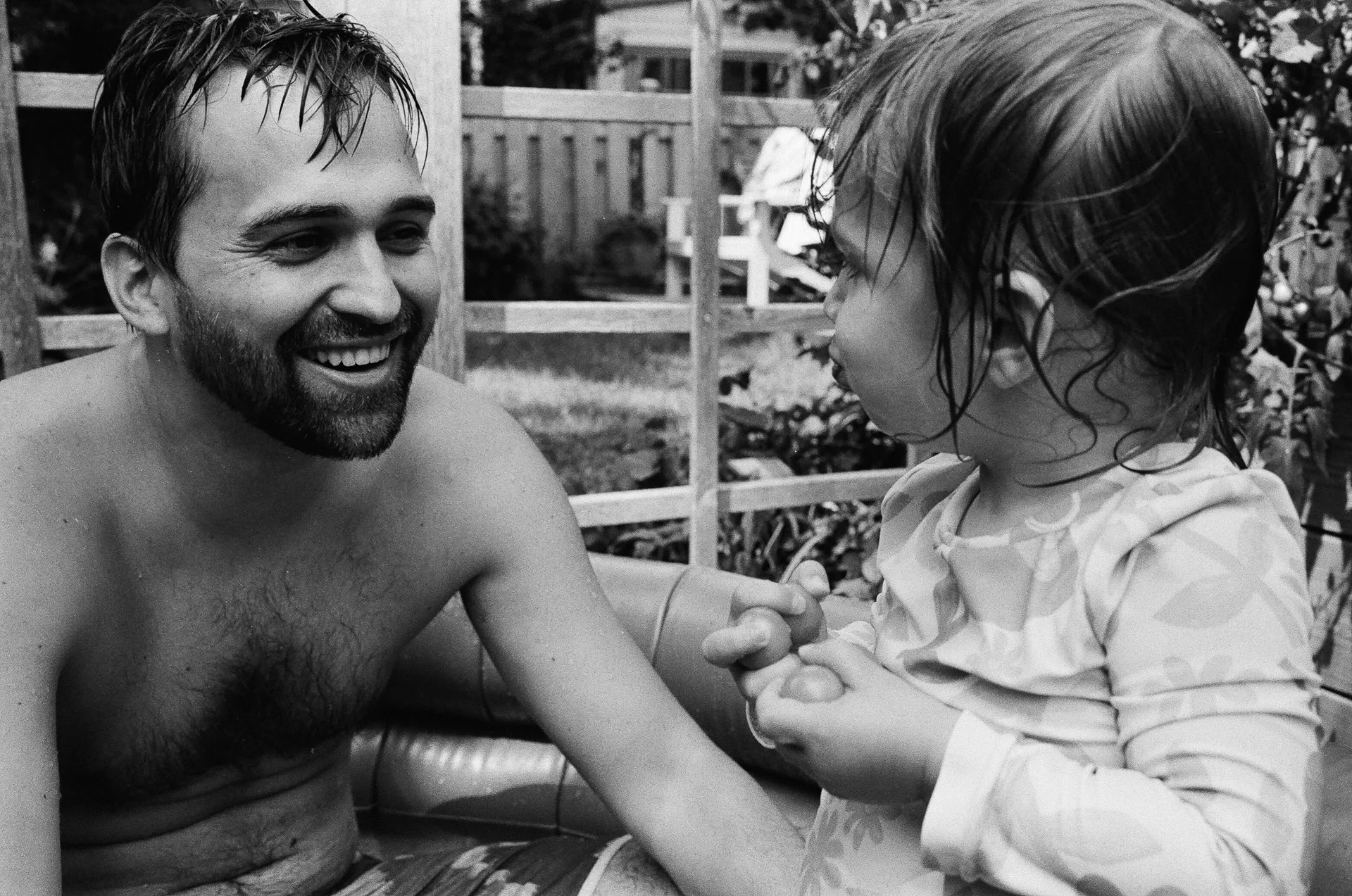 A man with a beard smiling while in a swimming pool, talking to a young girl with wet hair and a patterned shirt, holding her hands, outdoors in a backyard.