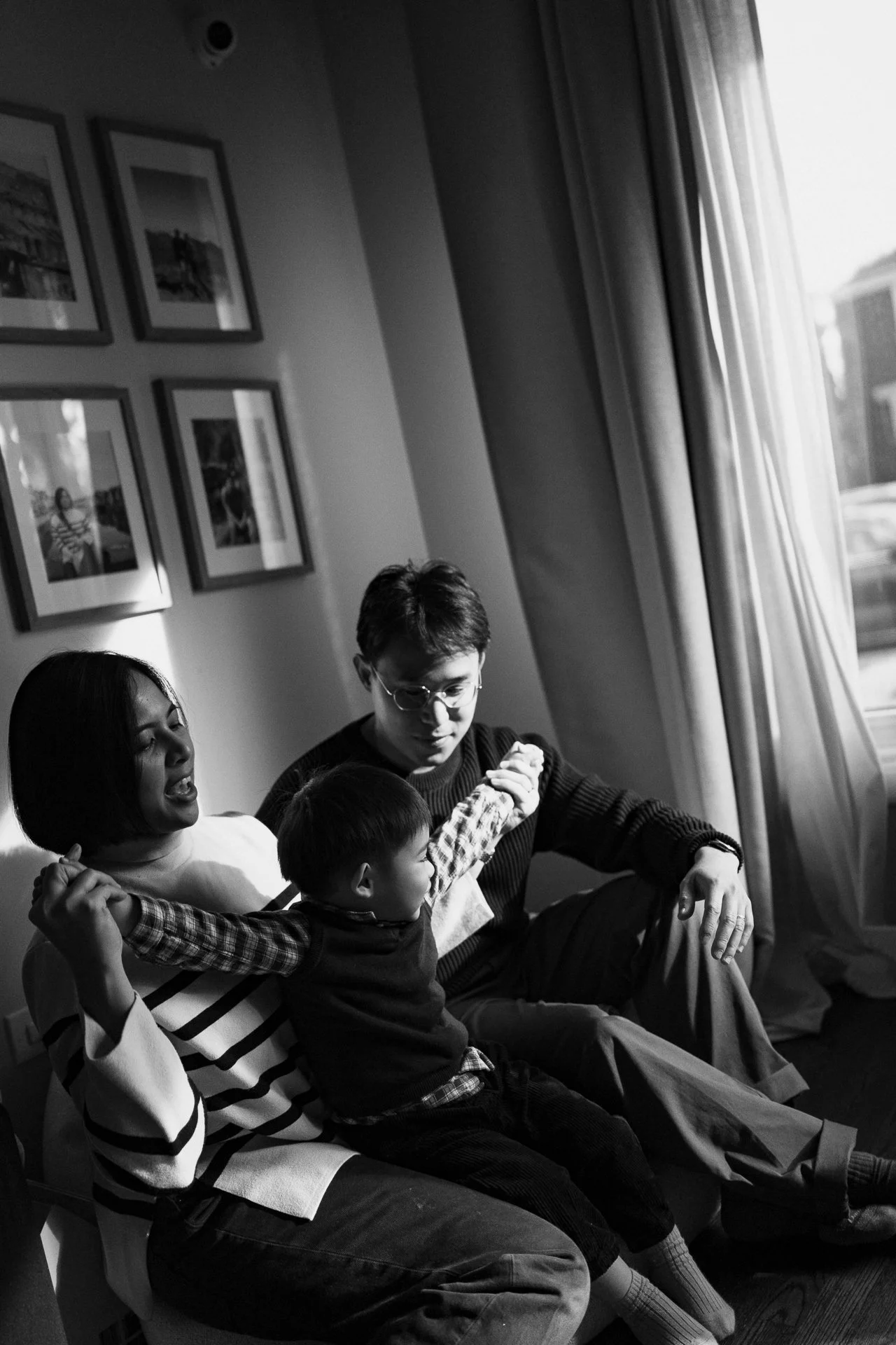 Black and white photo of three people sitting on a couch. A woman and a man are holding a young boy, who is stretching his arms. They are near a large window with curtains, and framed pictures are on the wall behind them. The woman's mouth is open, p