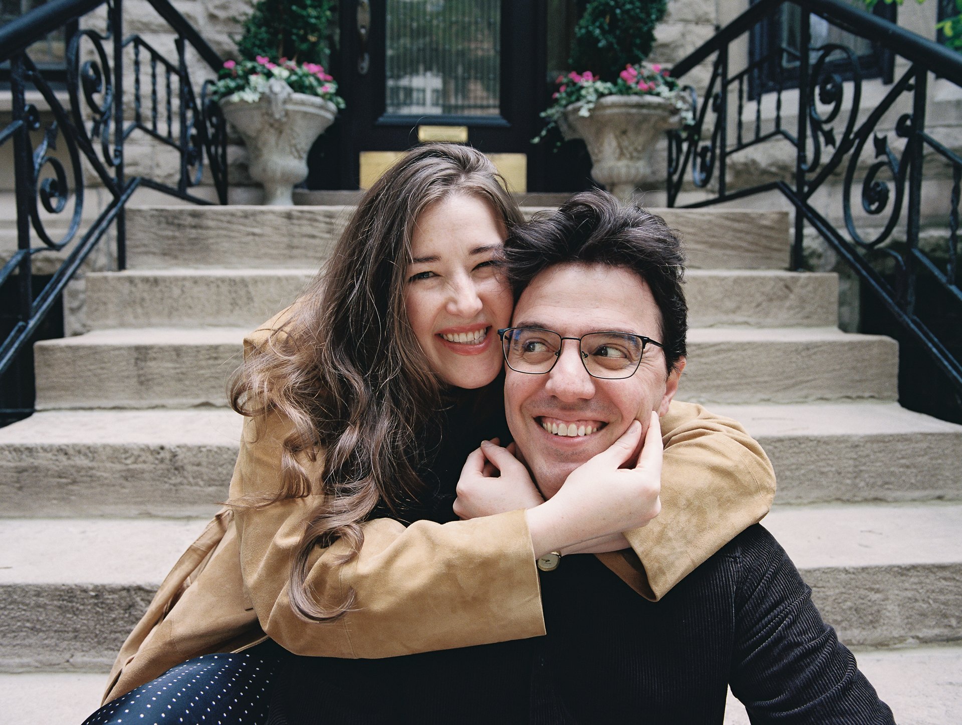 A happy young woman with long wavy brown hair embracing a smiling young man with glasses on a stone staircase outside a building. Two large planters with pink and white flowers are visible in the background.