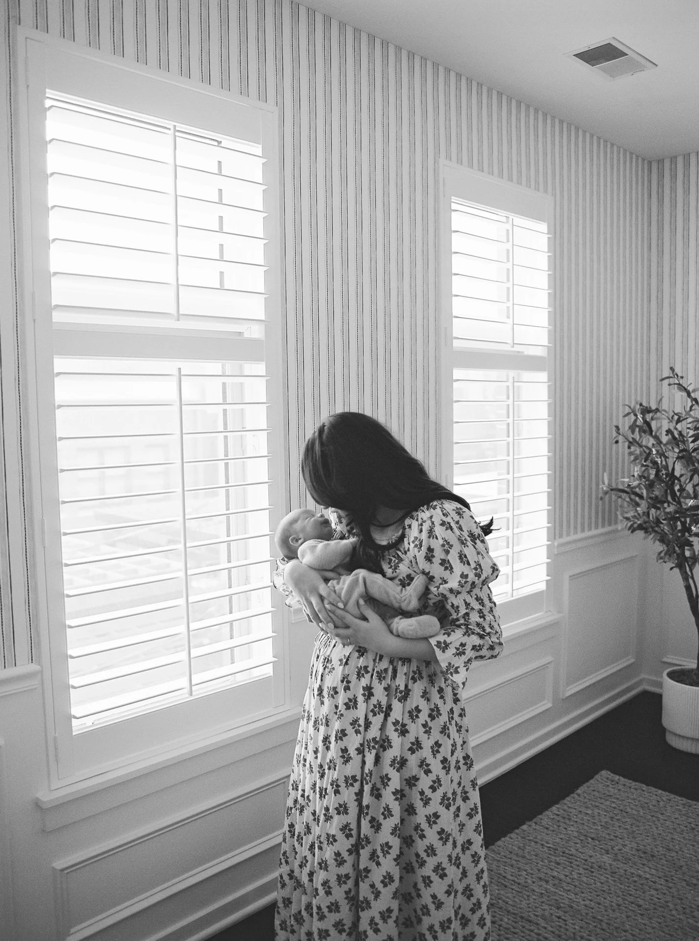 A woman holding a baby inside a room with two large windows with blinds and a potted plant.