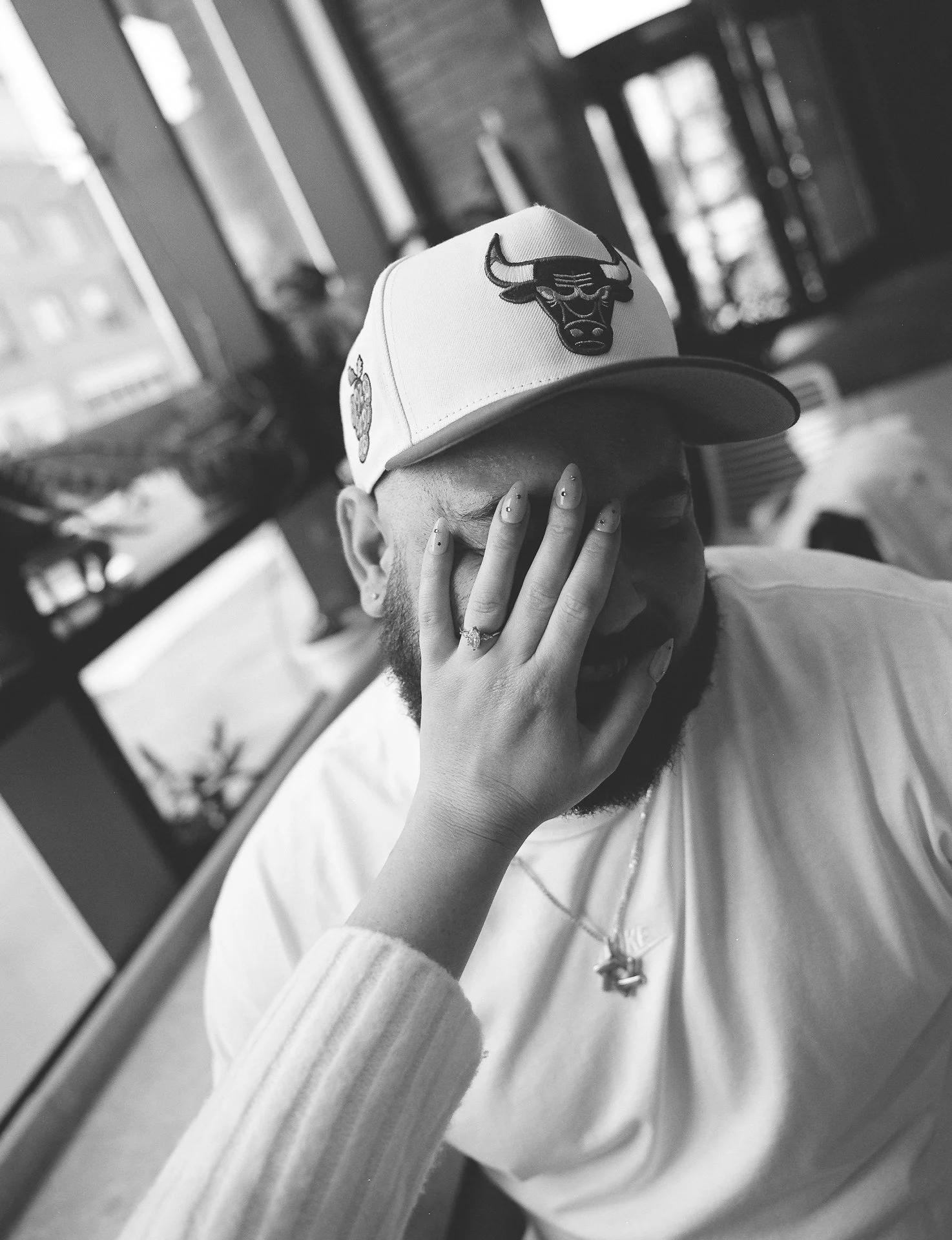 A man with a beard wearing a Chicago Bulls cap, covering his face with a woman's hand, laughing in a casual setting.