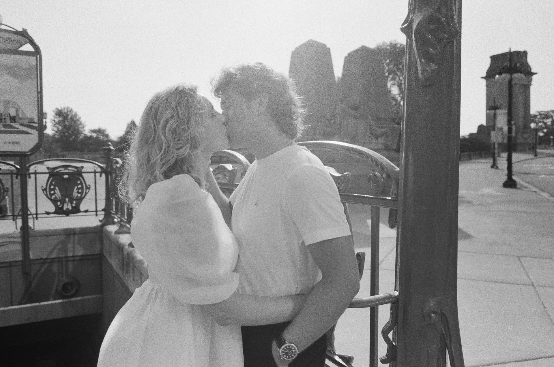 A young couple sharing a kiss in front of a carousel with tall buildings in the background.