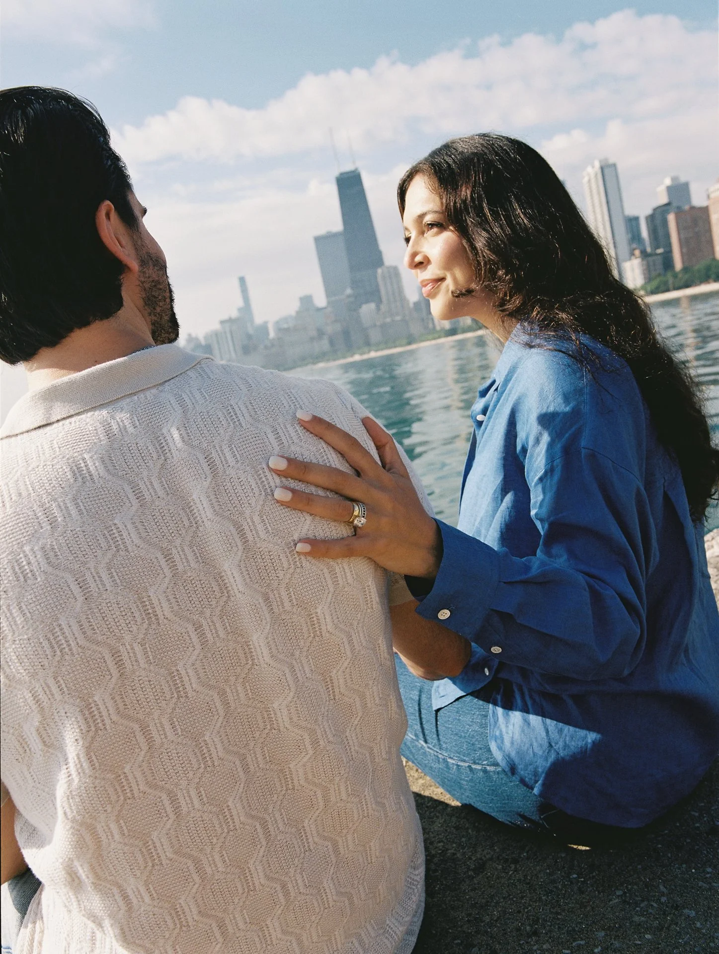 Woman sitting near a river with a city skyline in the background, looking at a man who is facing away from the camera.