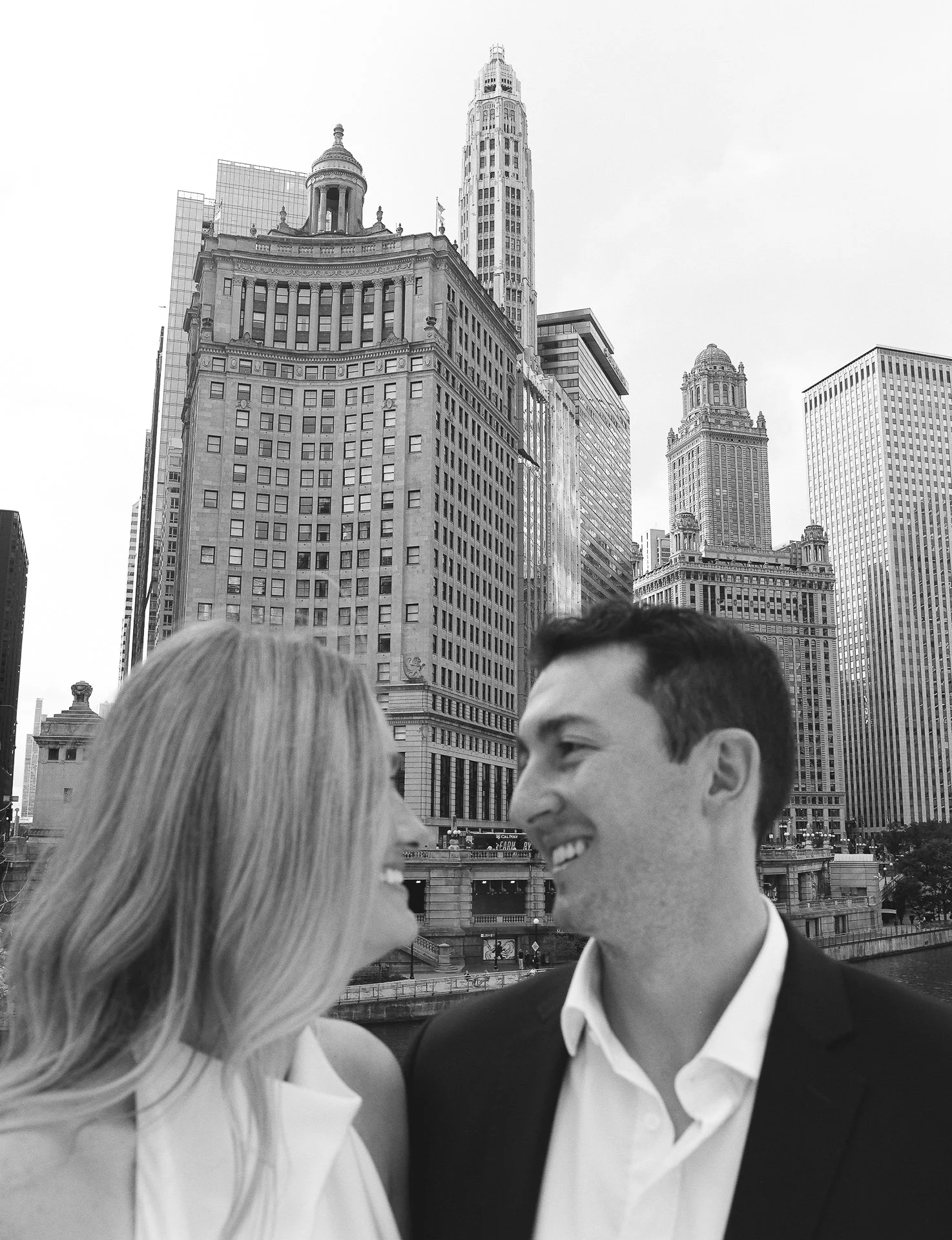 A black-and-white photo of a smiling couple looking at each other with a city skyline featuring tall buildings in the background.