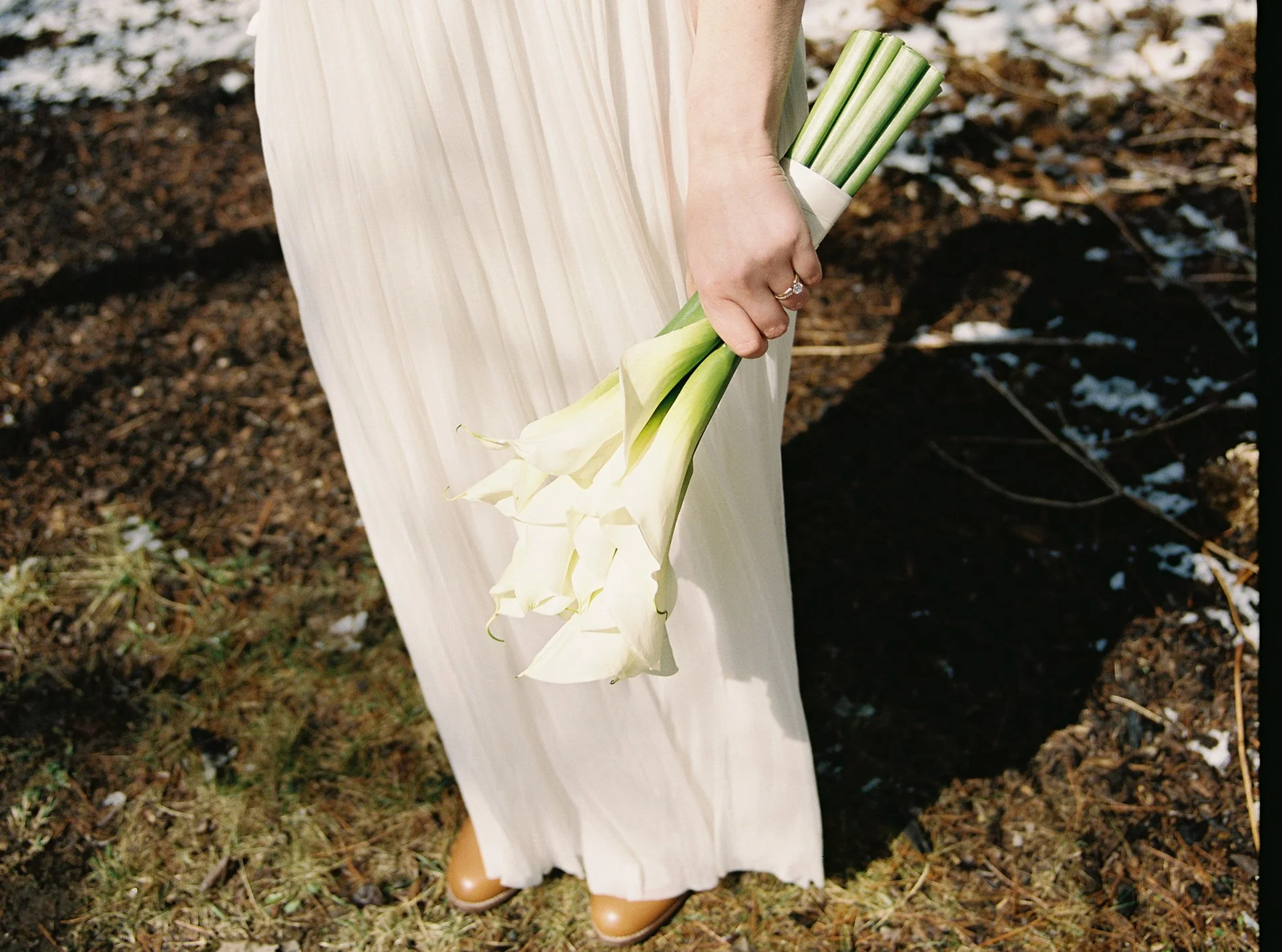 A person wearing a cream-colored dress and tan boots holding a bouquet of white calla lilies outdoors.