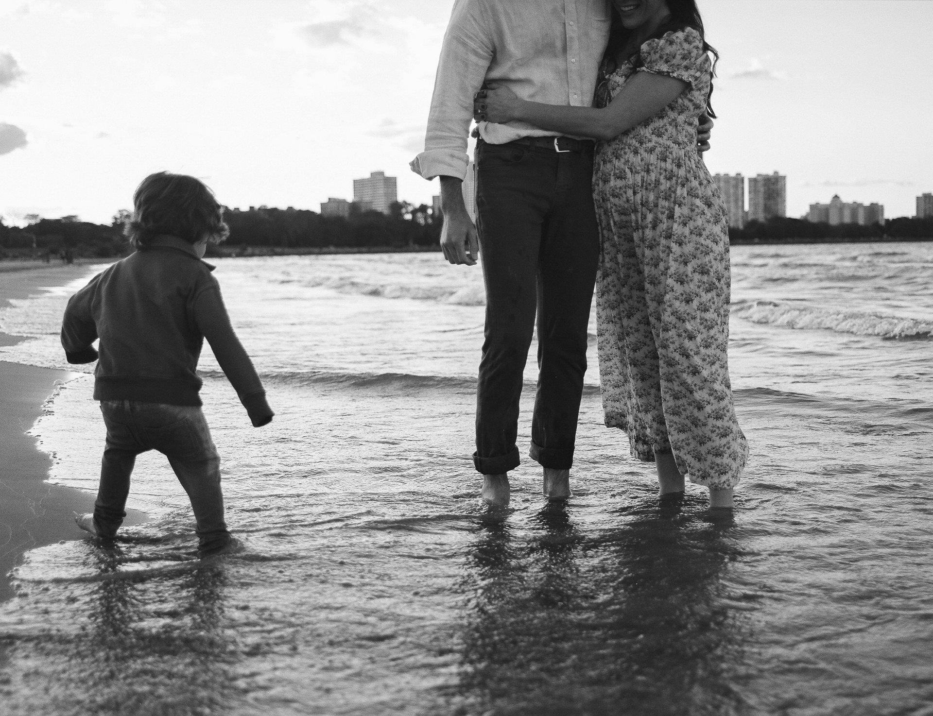 A black and white photo of a family at the beach, showing a man and woman holding each other and a small child walking in the shallow water, with city buildings in the background.